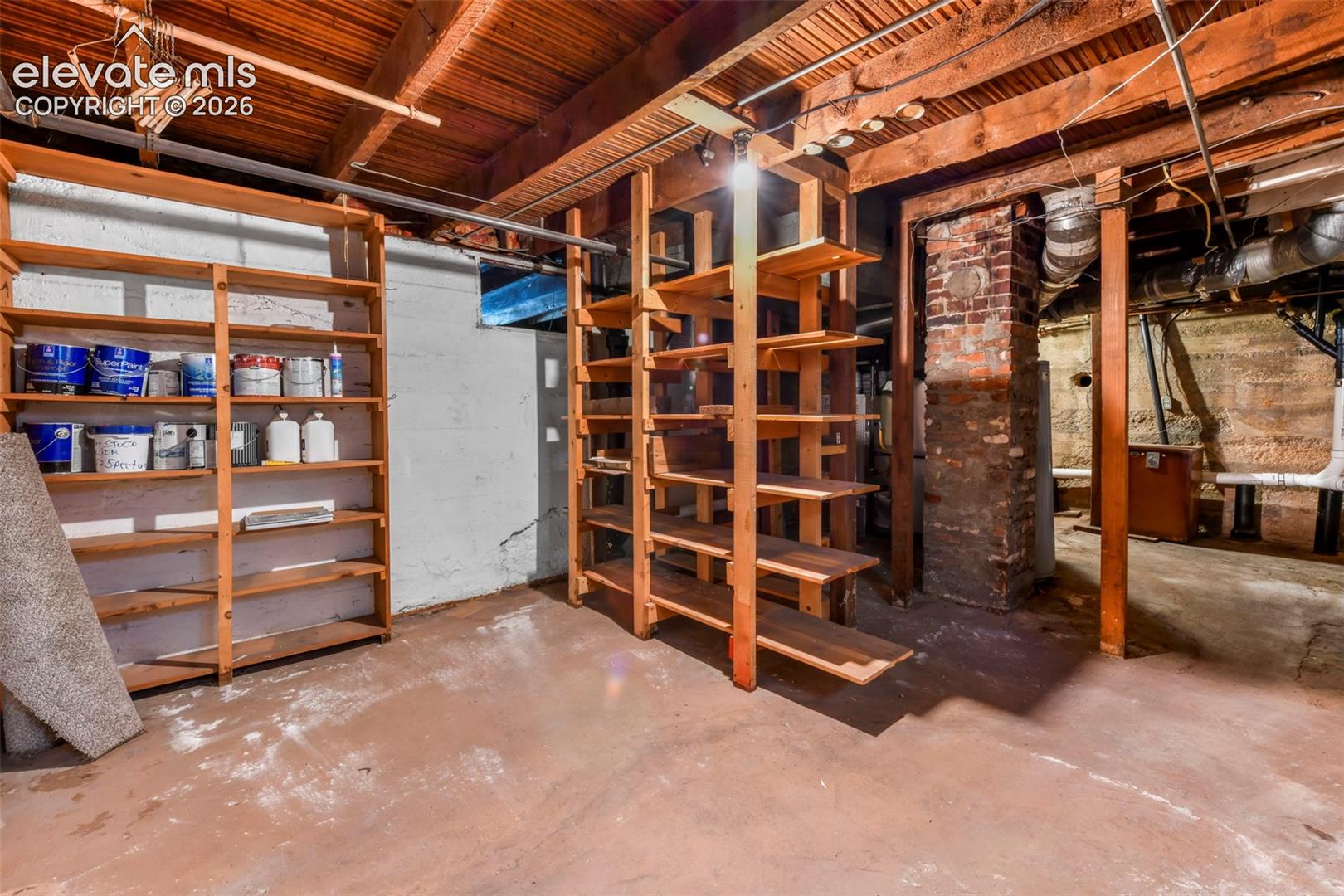 Basement storage area featuring exposed wood ceiling joists, masonry walls, a brick chimney, and floor-to-ceiling wooden shelving units