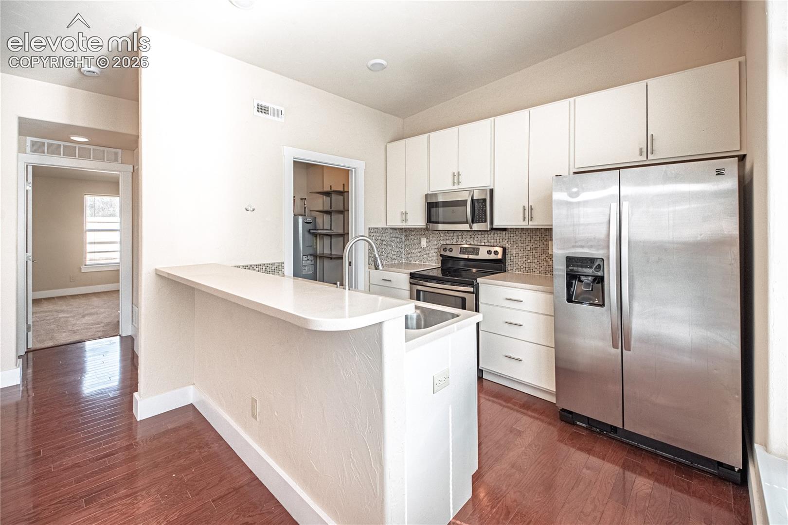 Kitchen featuring wood-finish flooring, white cabinetry, stainless steel appliances, mosaic tile backsplash, and a breakfast bar with a solid surface countertop