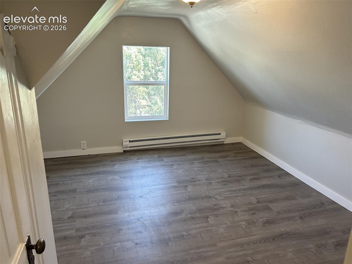 Attic room featuring wood-finish flooring, an angled ceiling, a single window, and a baseboard heater