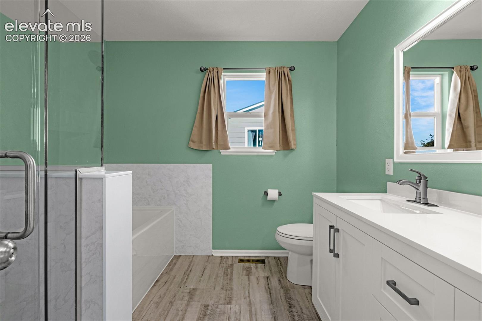 Bathroom featuring a vanity with a white countertop, wood-finish flooring, a glass-enclosed shower, a deep soaking tub with a white surround, and a framed mirror