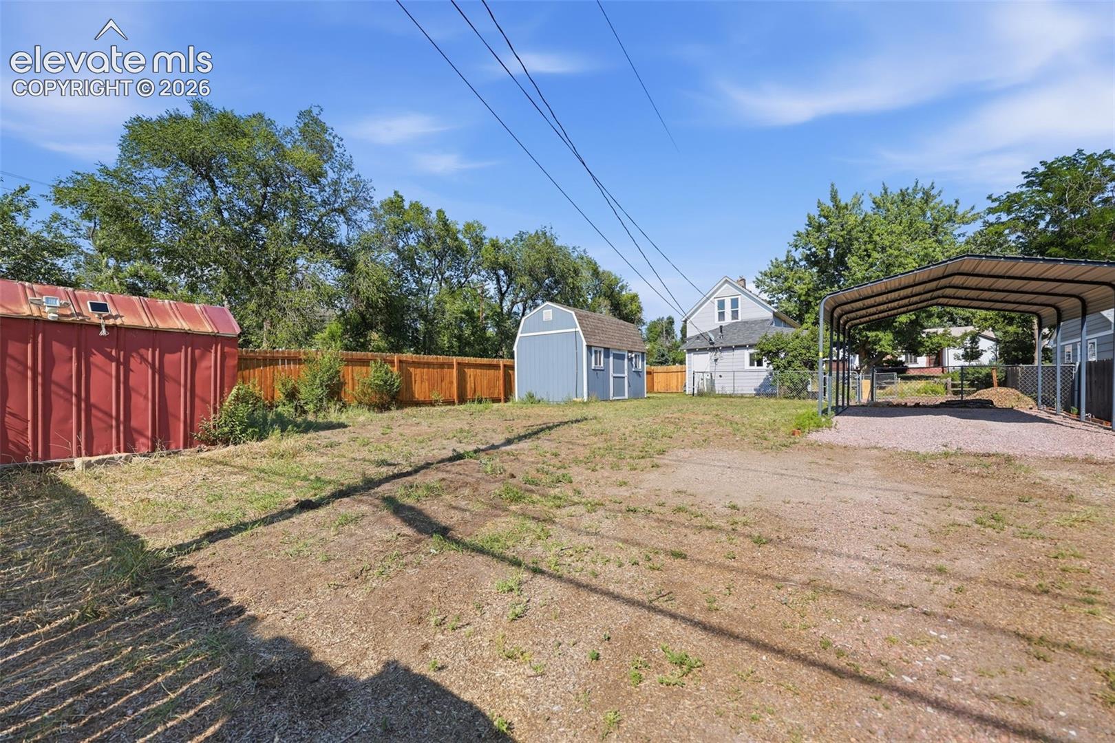 Expansive property featuring a red storage shed, blue storage shed, wood privacy fencing, and a metal carport.
