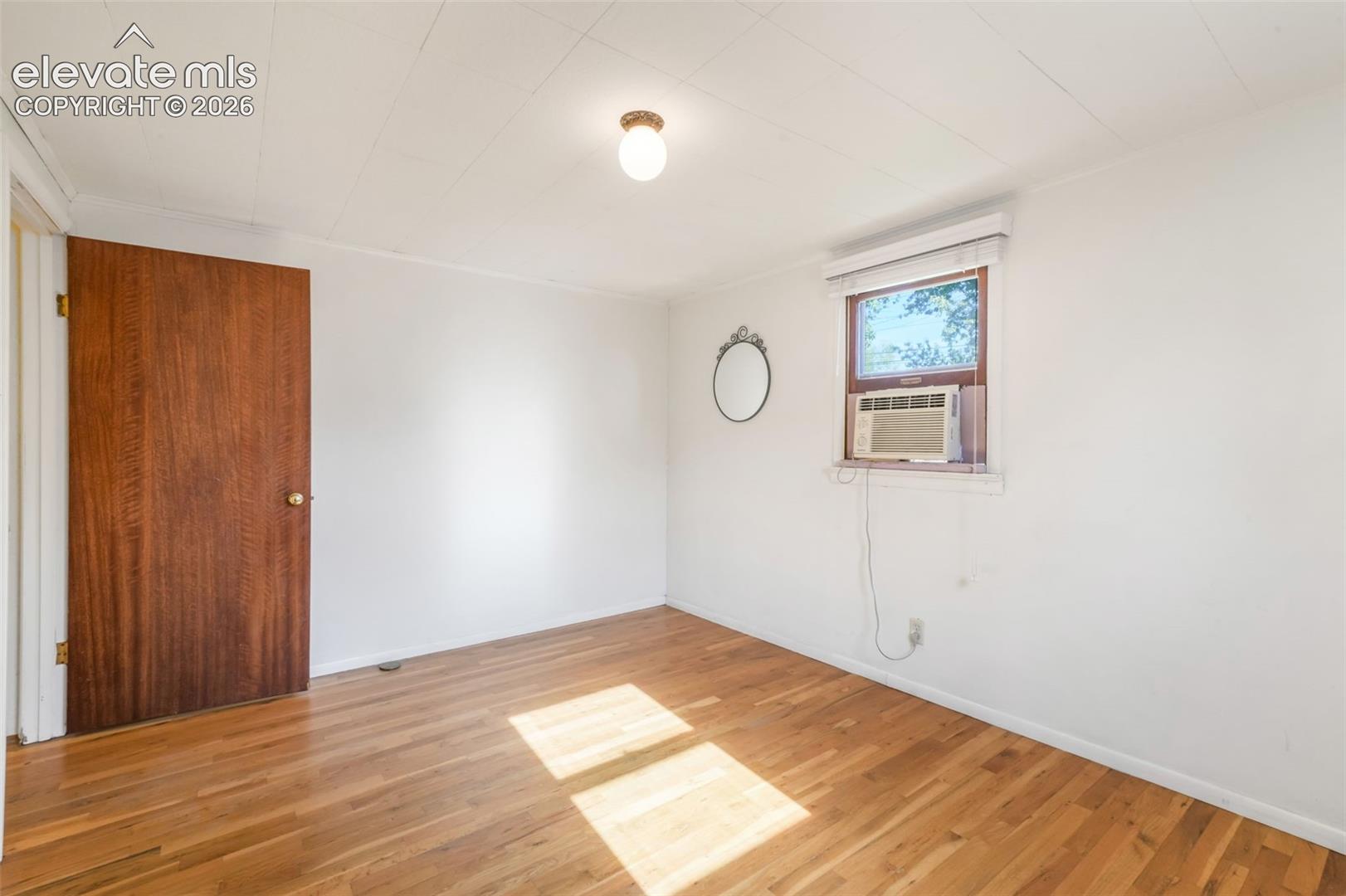 Hardwood flooring, white painted walls, window with blinds, ceiling-mounted light fixture, and a wood-finish door