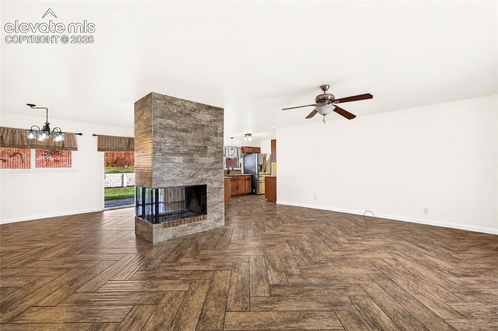 Open-concept living space featuring a central, stone-tiled dual-sided fireplace, herringbone-pattern wood-finish flooring, a ceiling fan with integrated lighting, and a multi-arm chandelier