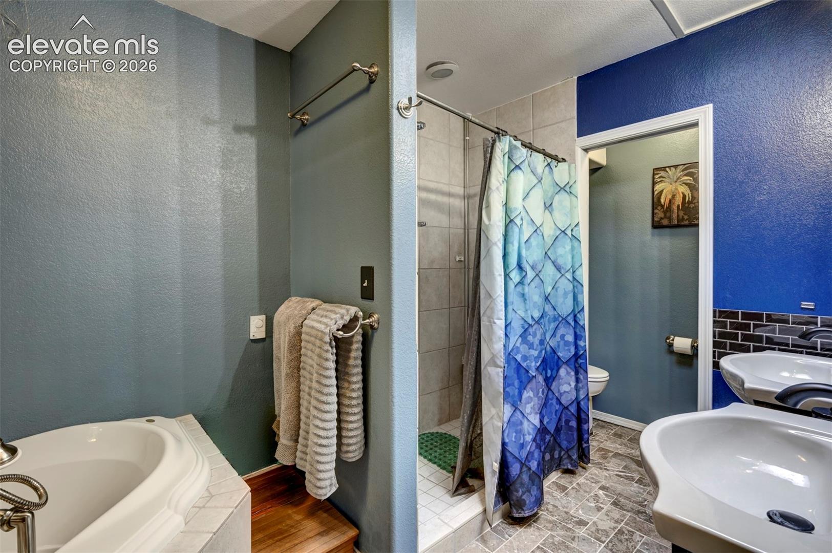 Built-in soaking tub with tiled surround, walk-in shower with tiled walls, and dual pedestal sinks