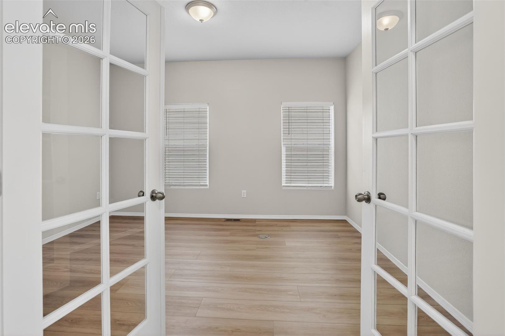 Double French doors with frosted glass panels open into a room featuring wood-finish flooring, light grey walls, and two windows with horizontal blinds
