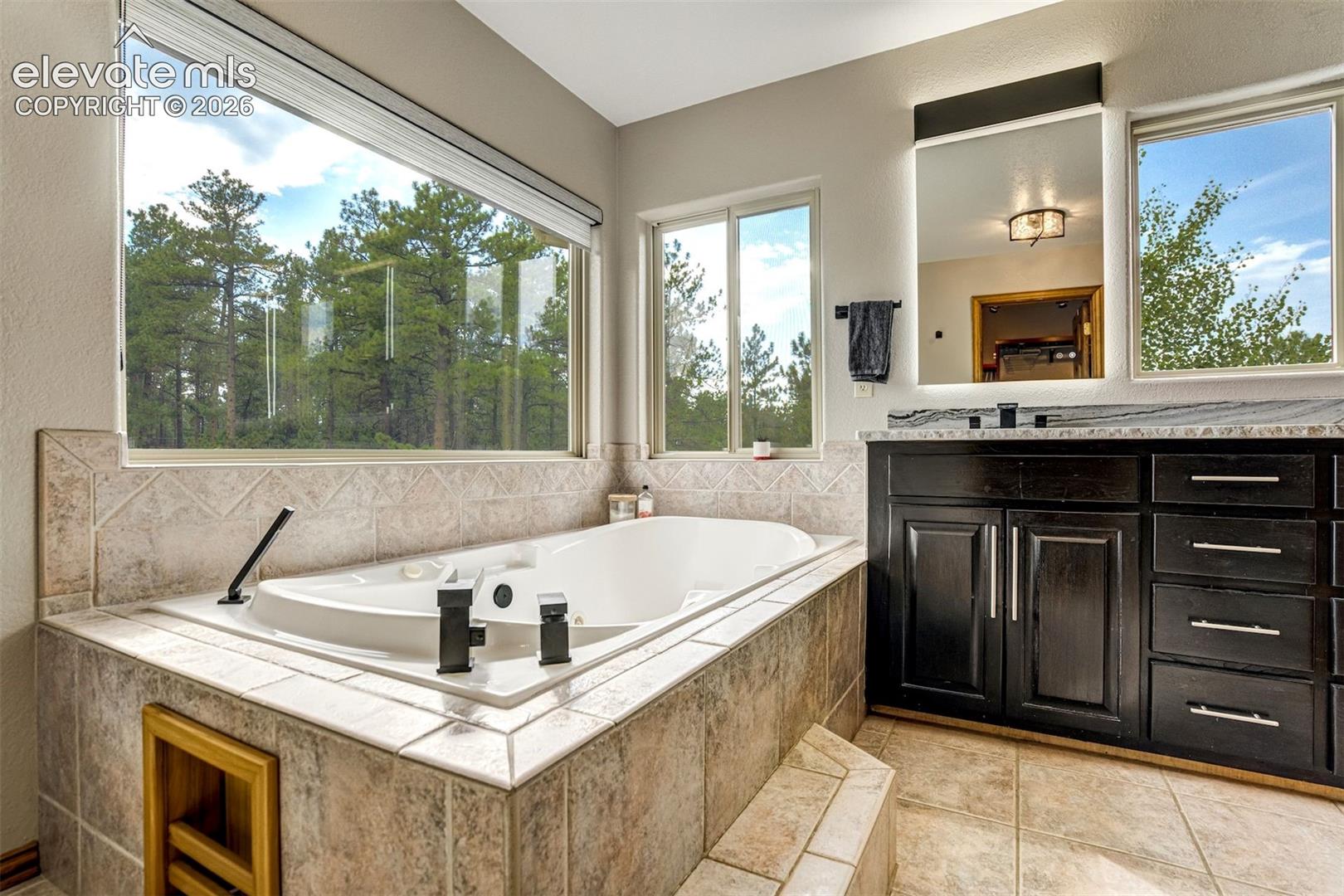 Soaking tub with tile surround, expansive windows, dark wood-finish vanity with stone countertop, and an illuminated mirror