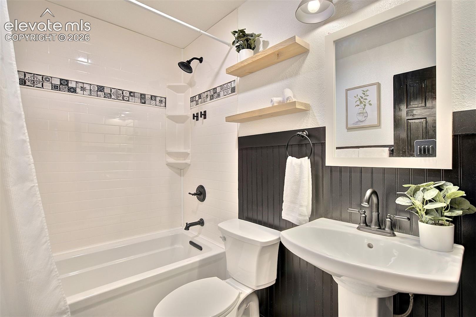 Bathroom featuring a white pedestal sink with dark metallic hardware, a white subway-tiled shower surround with decorative accent tiling, black beadboard wainscoting, and natural wood-finish wall-mounted shelving