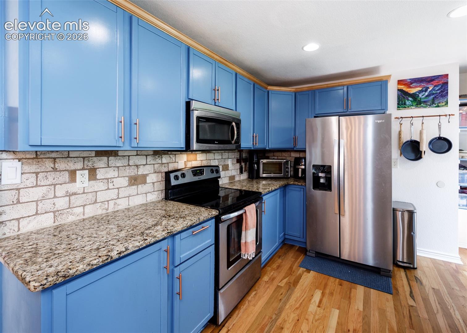Blue cabinetry with gold-tone hardware, granite countertops, brick-style tile backsplash, stainless steel appliances, and wood-finish flooring