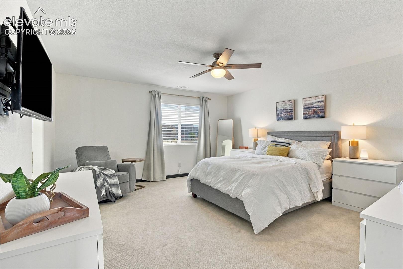 Wall-mounted television with articulating bracket, ceiling fan with integrated lighting, window with blinds and drapery, and neutral carpeting