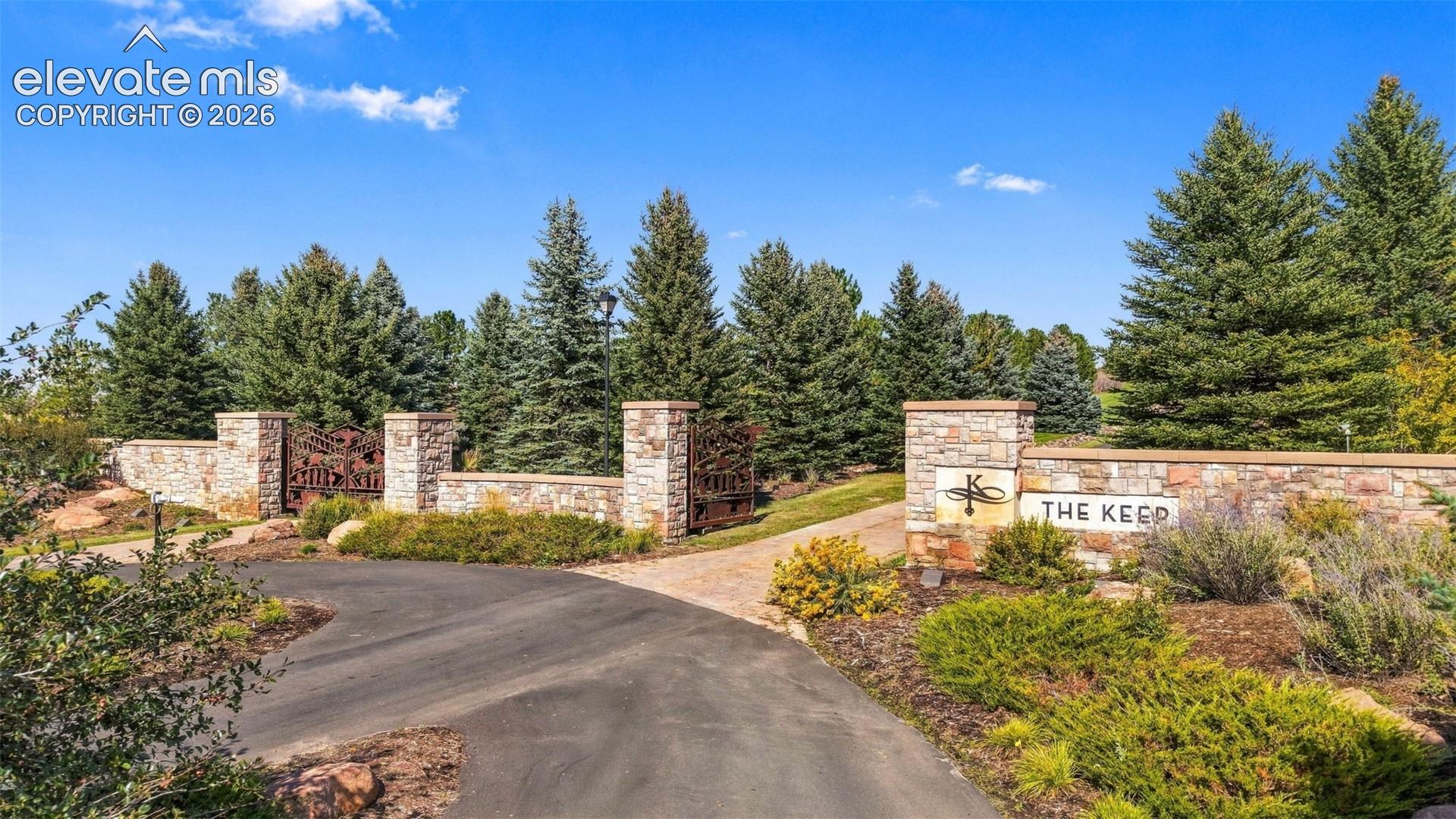 Stone entrance wall with integrated signage, wrought iron gates, mature evergreen trees, and paved driveway
