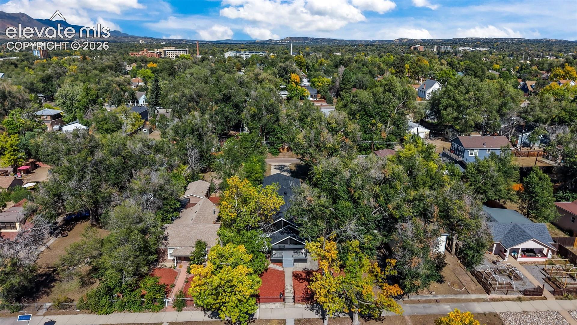 Aerial view showcasing a substantial tree-lined lot with mature landscaping, red brick residence with dark roof, detached garage structure, and manicured grounds set against a backdrop of rolling foothills and established neighborhood canopy
