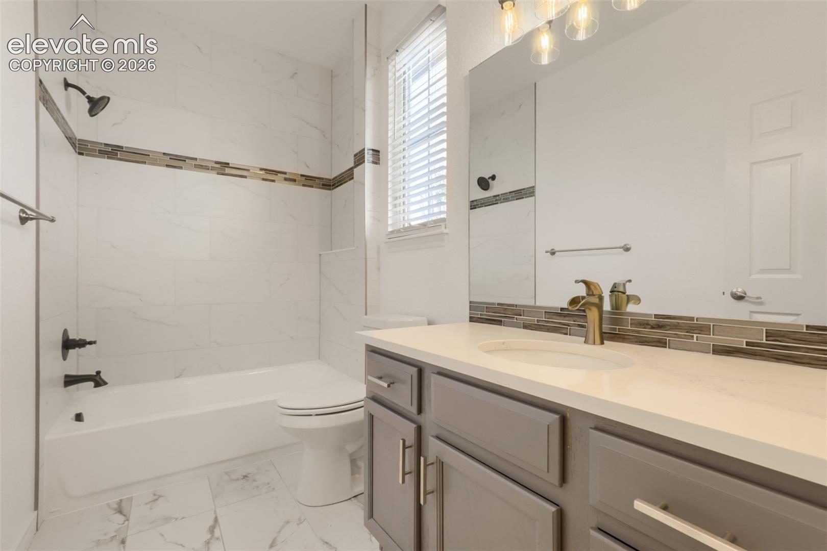 Stylishly updated second-floor full bathroom showcasing modern grey cabinetry, an undermount sink, and bronze fixtures. Complete with a tub/shower combo surrounded by large marble-look tile, a mosaic accent strip, matching floor tile, and a bright window for natural light.