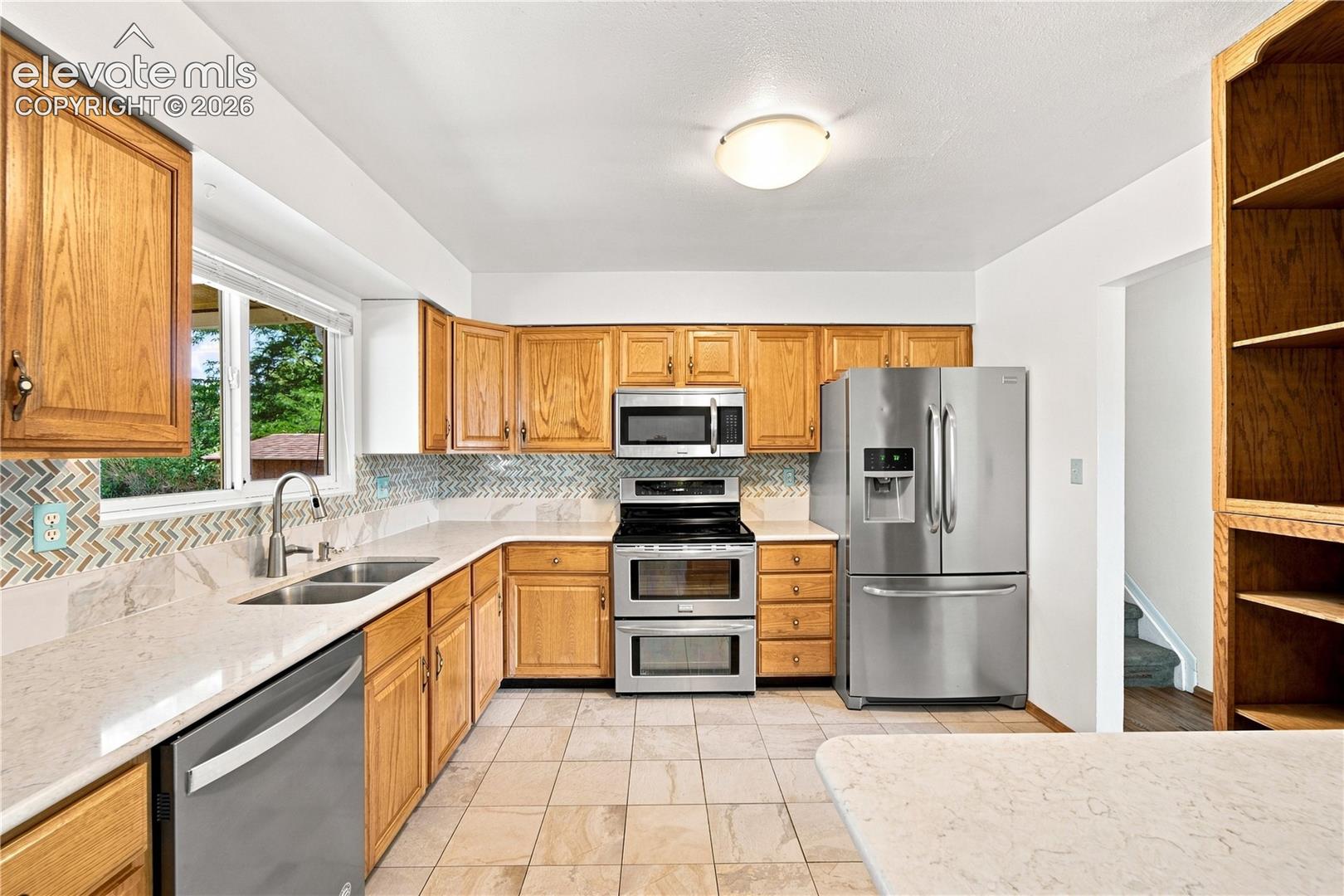 Kitchen featuring wood cabinetry, stainless steel appliances, a double basin sink, light-toned countertops, and mosaic tile backsplash