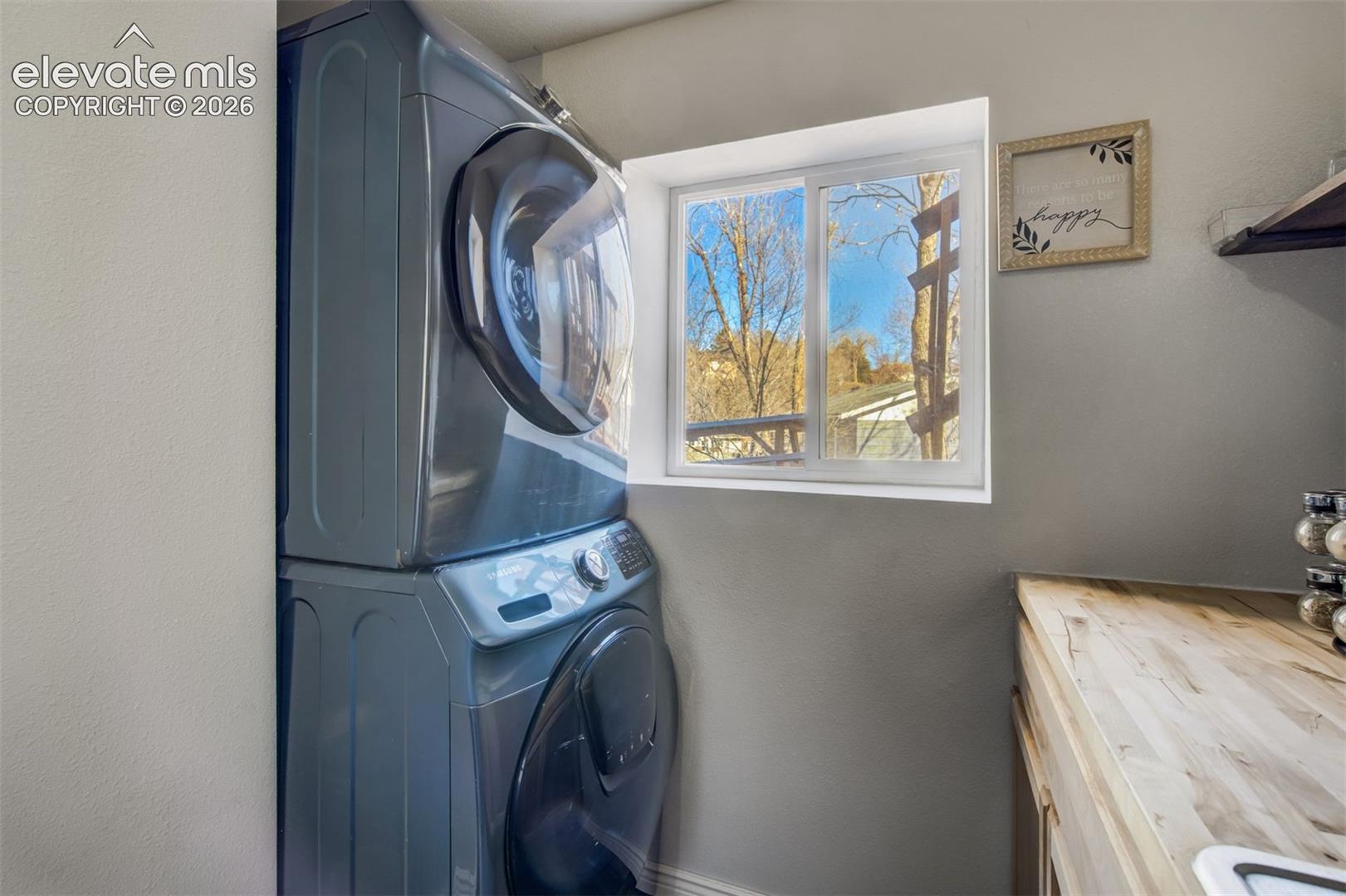 Stacked laundry appliances, wood-finish countertop, and a white-framed window