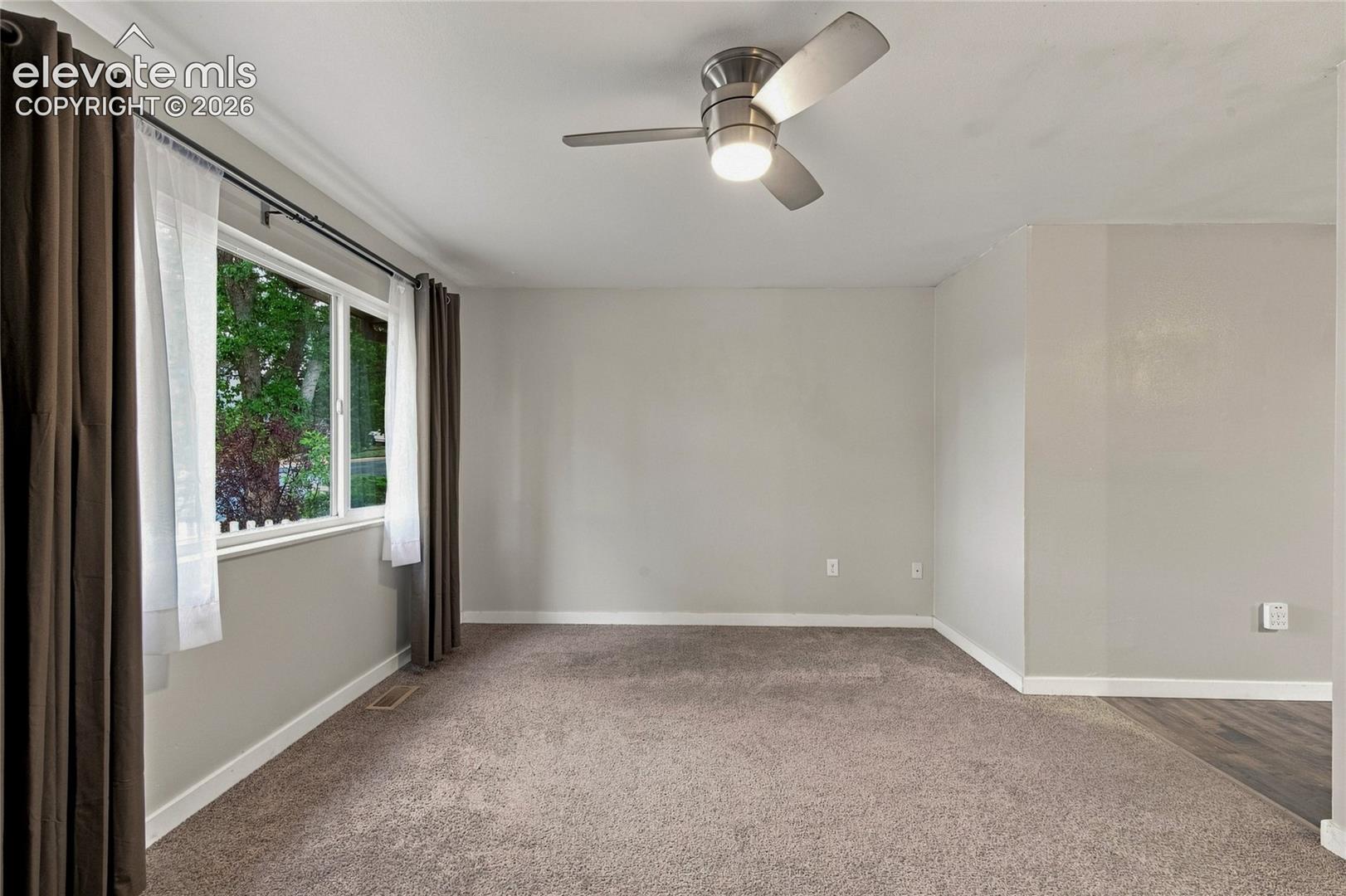 Bright, spacious living room upon entrance, featuring neutral wall finishes, plush carpet flooring, a ceiling fan with integrated lighting, and large windows with dual-layer curtain coverage allowing abundant natural light