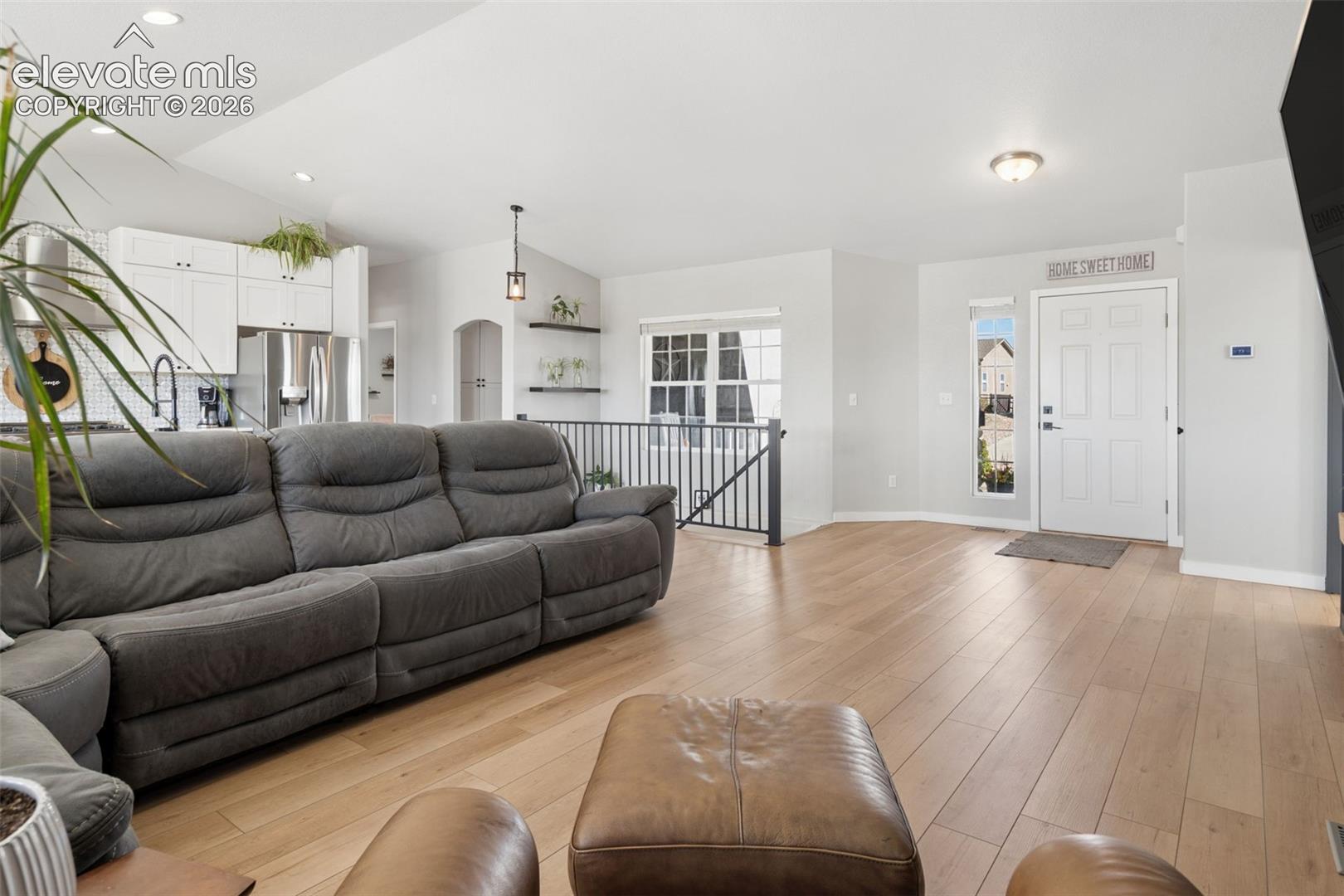 Vaulted ceiling, luxury vinyl flooring, white paneled entry door, kitchen with white cabinetry, and a black metal stair railing