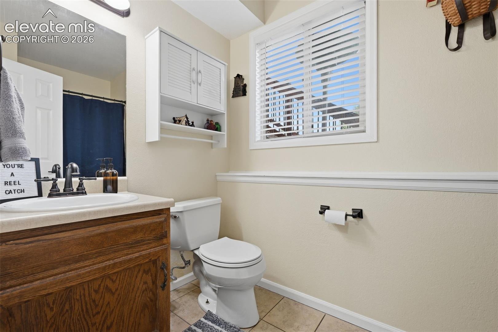 Bathroom featuring a wood-finish vanity with a built-in sink, white cabinetry with louvered doors, a window with horizontal blinds, and a toilet