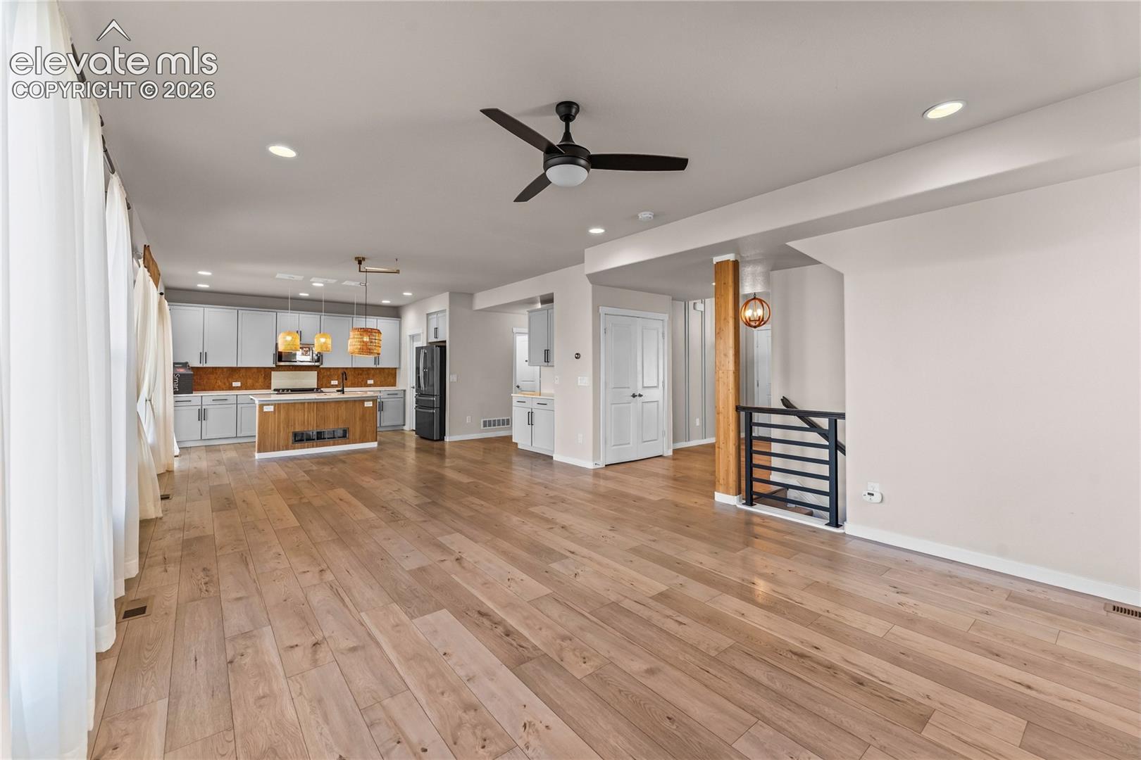 Open-concept interior featuring wood-finish flooring, recessed lighting, matte black ceiling fan, wood-accented center island, and metal-railing staircase