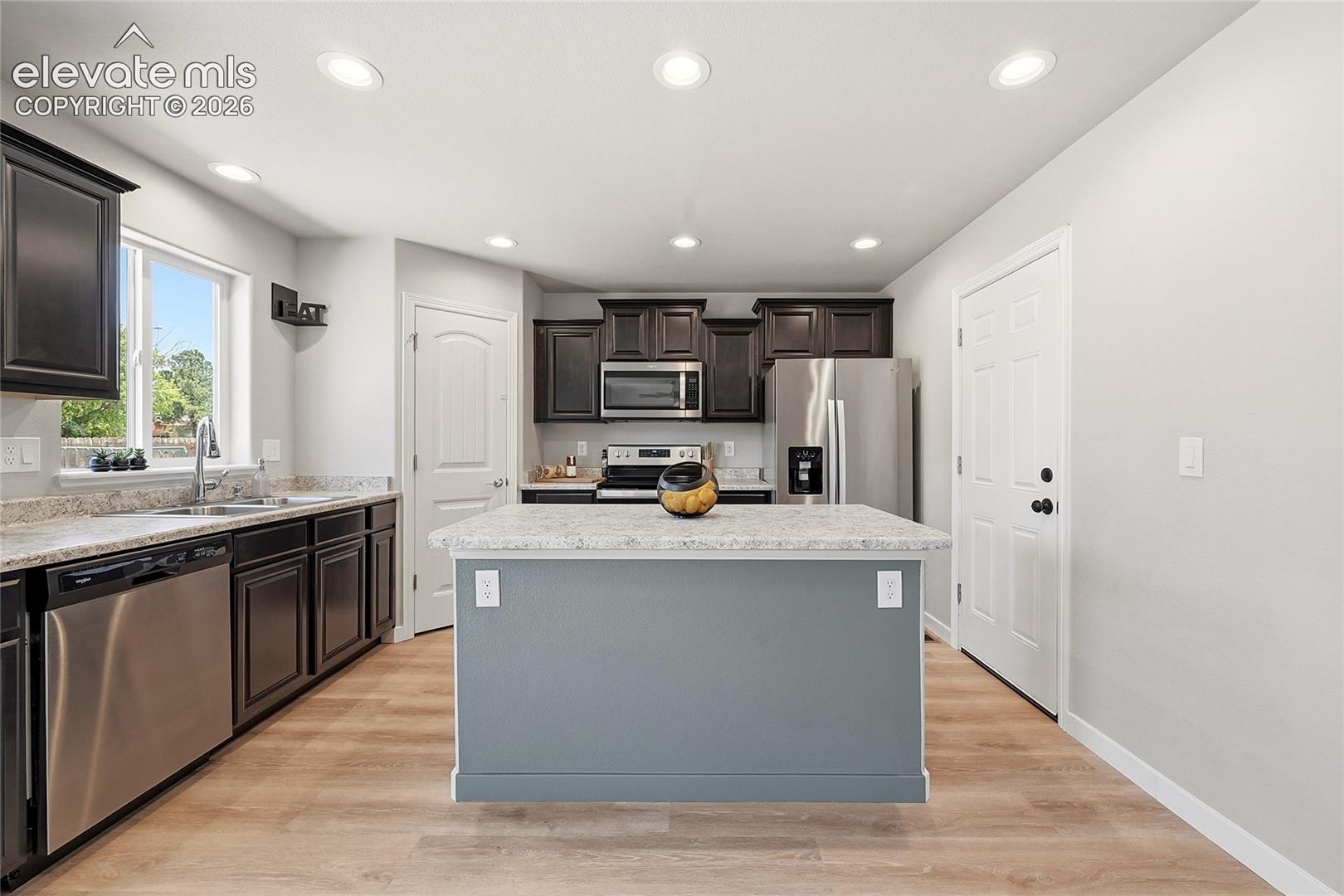 Kitchen featuring a central island with a light-toned countertop, dark wood-finish cabinetry, stainless steel appliances, recessed lighting, and wood-finish flooring