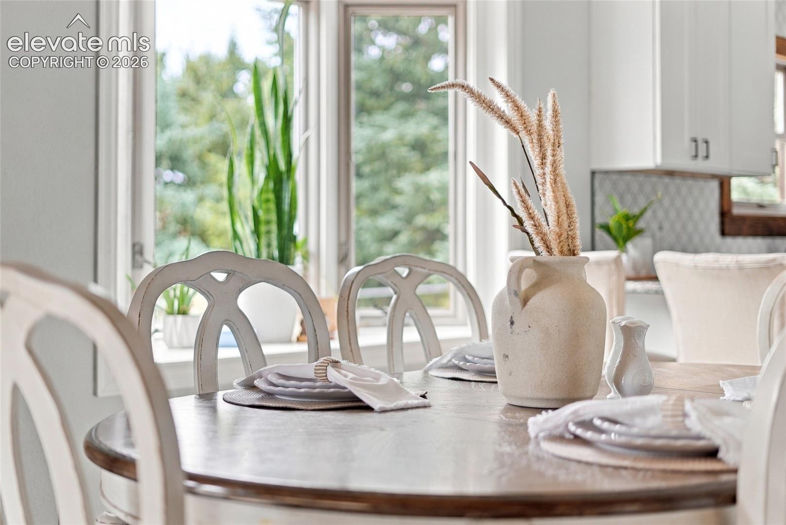 Wood-finish dining table with light-toned chairs, abundant natural light, and a tiled backsplash in the background