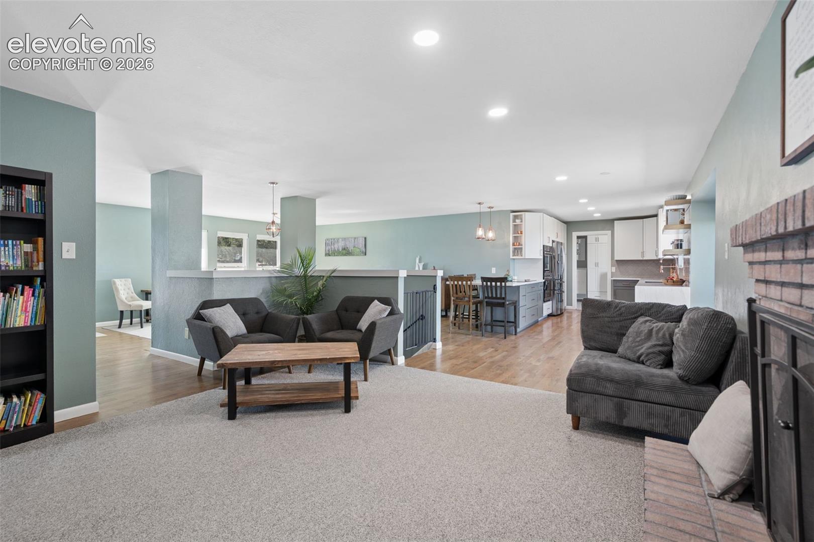 Open living area featuring wood-finish flooring, recessed lighting, a brick fireplace, built-in shelving, and a kitchen island with pendant lighting