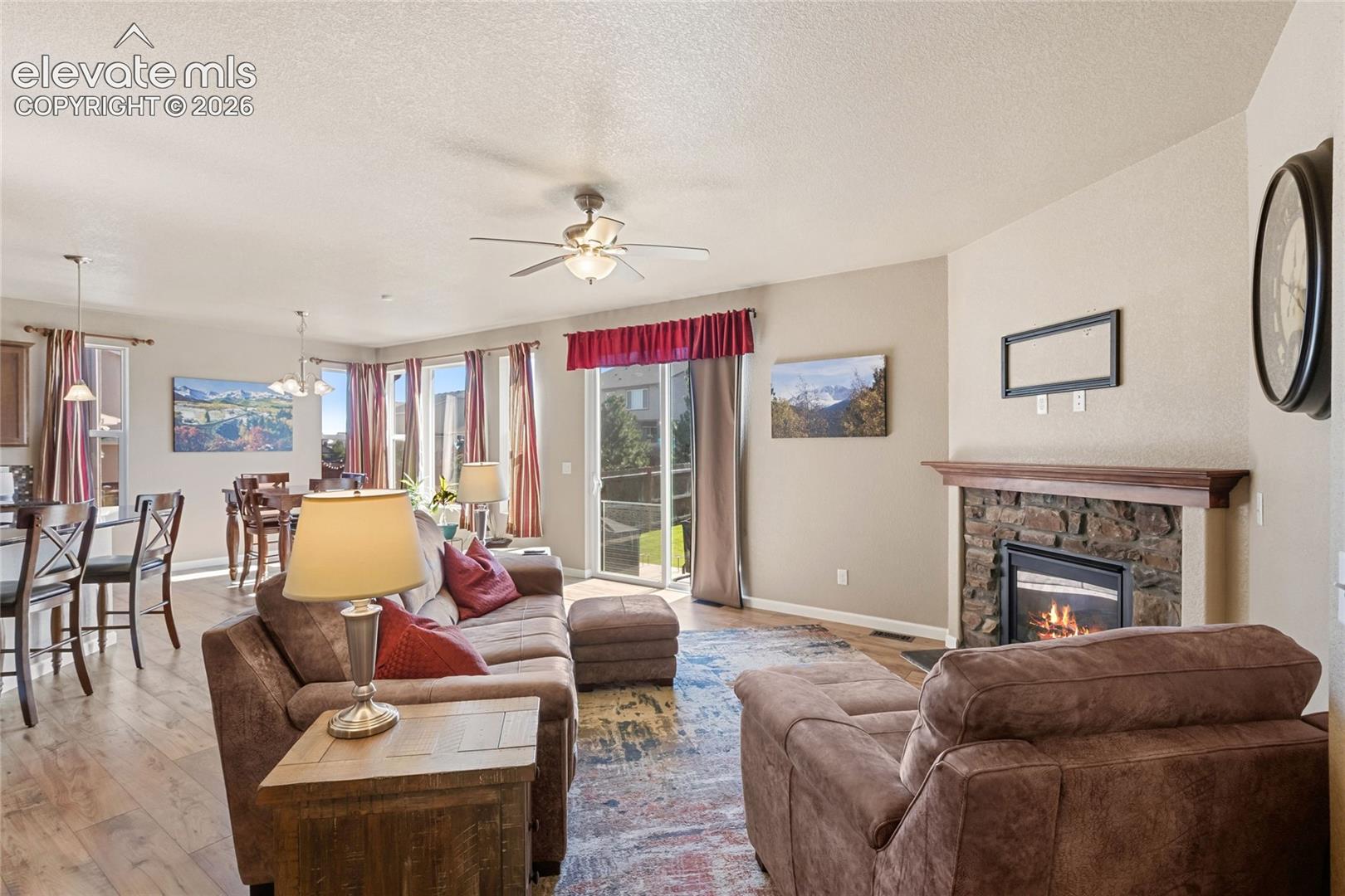 Open-concept interior featuring wood-finish flooring, a stone-surround fireplace with a timber mantel, a ceiling fan, and sliding glass doors