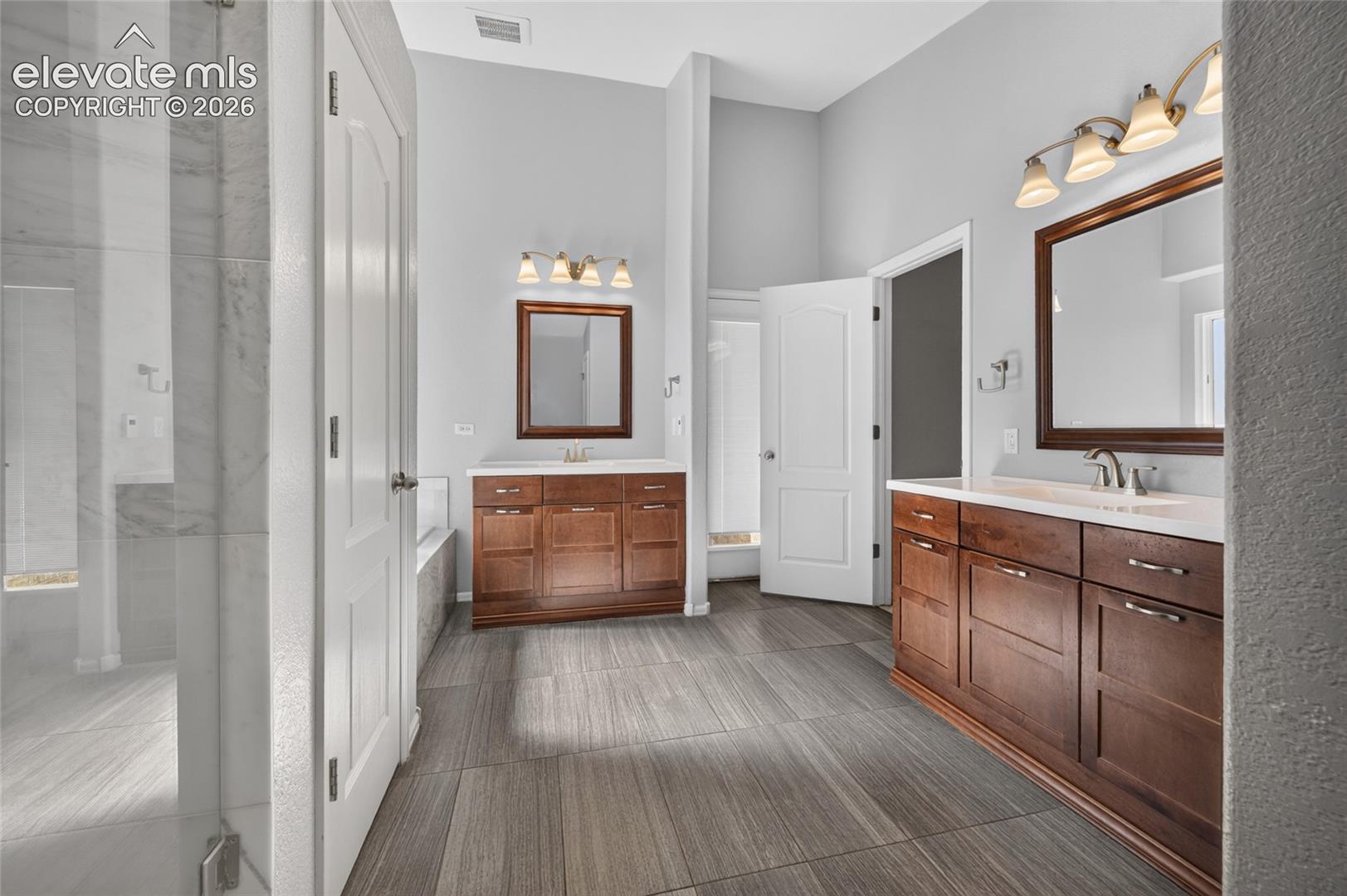 Dual wood-finish vanities with light-toned countertops, framed mirrors, high-output vanity lighting, and ceramic tile flooring