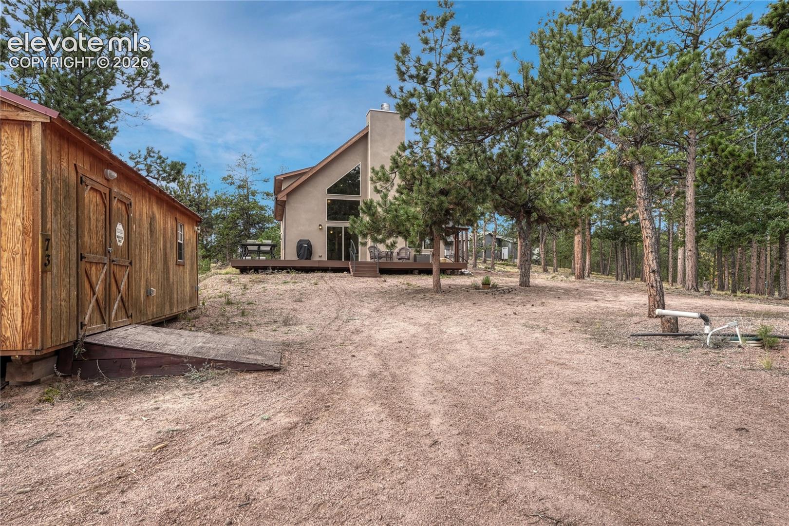 Structure with stucco exterior, floor-to-ceiling windows, attached  deck, and detached wooden storage structure (workshop) on a treed lot