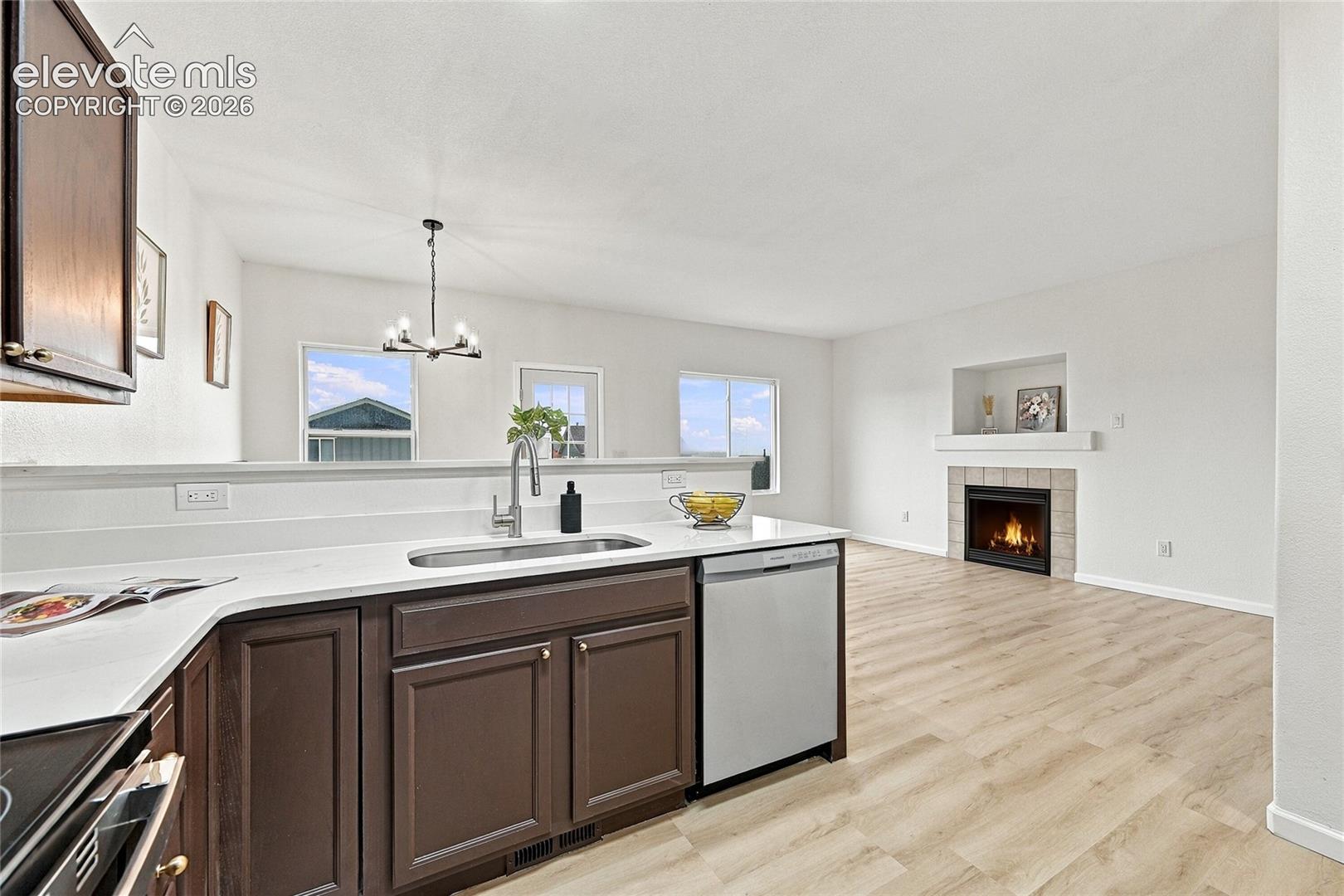 Kitchen featuring dark-toned cabinetry, solid-surface countertops, an undermount sink, and stainless steel dishwasher, complemented by wood-finish flooring and a fireplace with a mantel