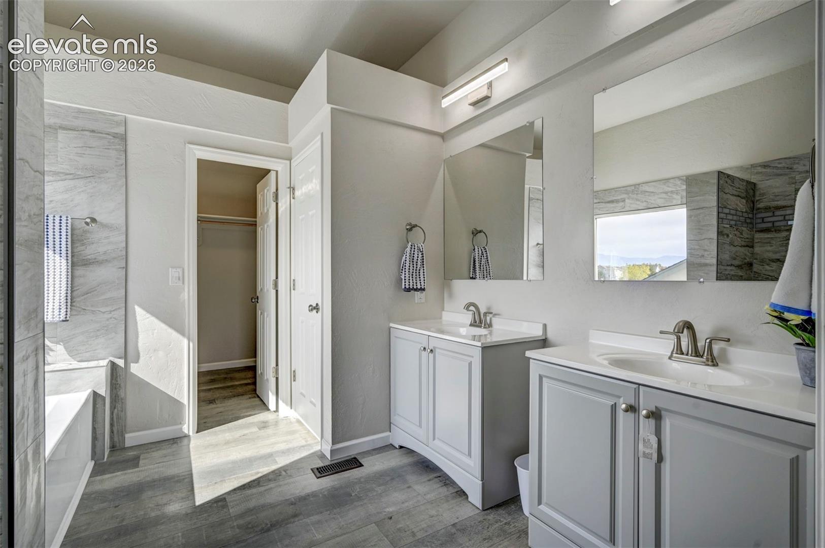 Dual vanities with shaker-style cabinetry, white countertop surfaces, polished metal fixtures, wood-finish flooring, and wall-mounted vanity lighting