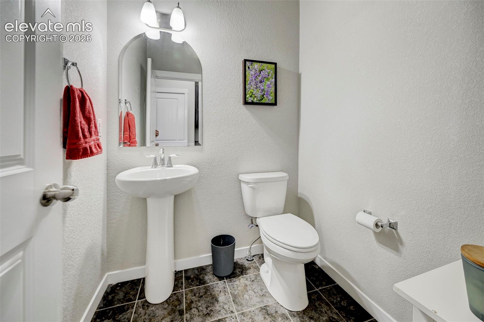 Pedestal sink with polished chrome faucet, arched wall mirror, two-light vanity fixture, and tiled flooring