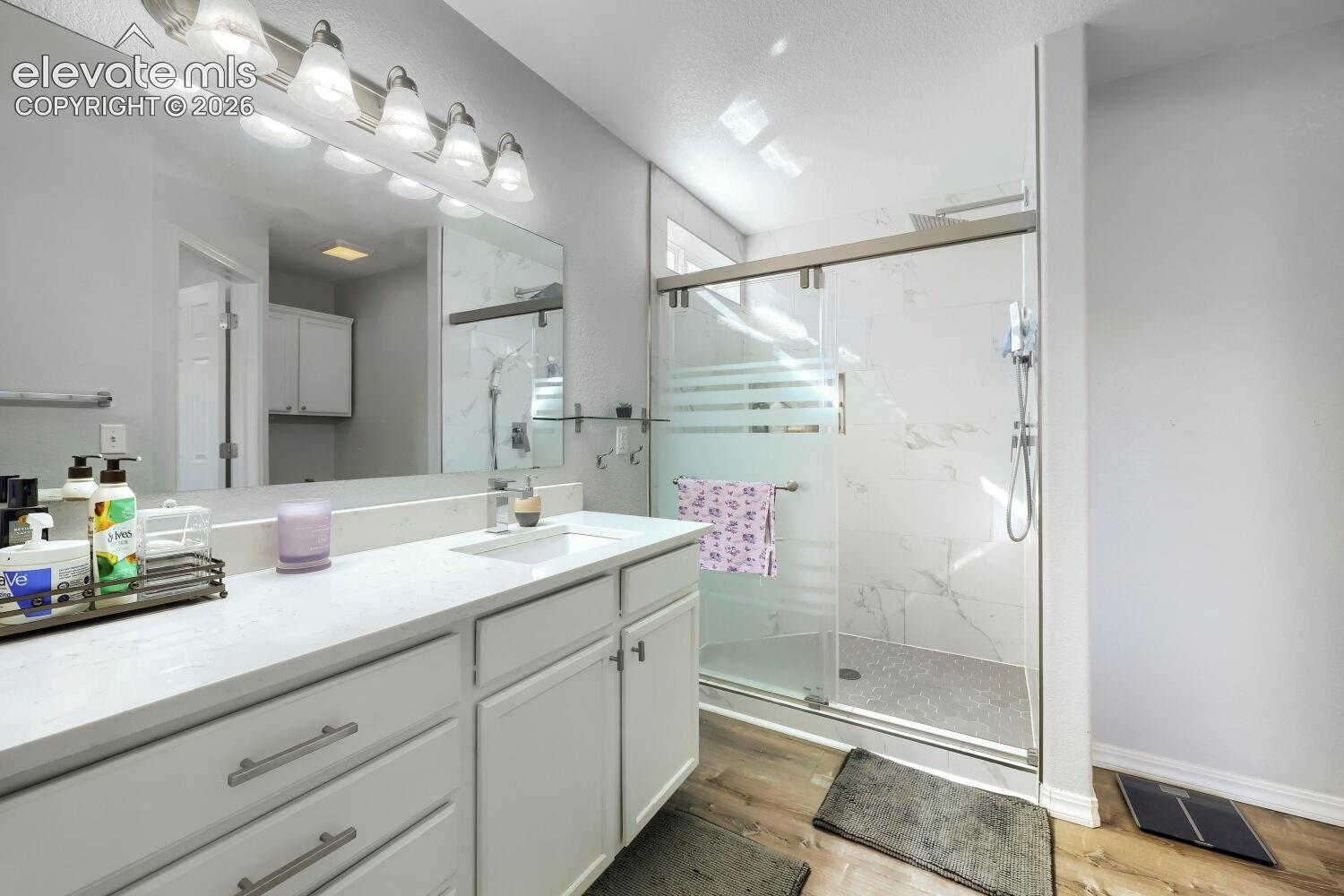 Bathroom featuring a white cabinetry vanity with a light-toned solid surface countertop, large wall-spanning mirror, multi-bulb vanity lighting, and a walk-in shower with sliding glass doors and stone-finish wall tile