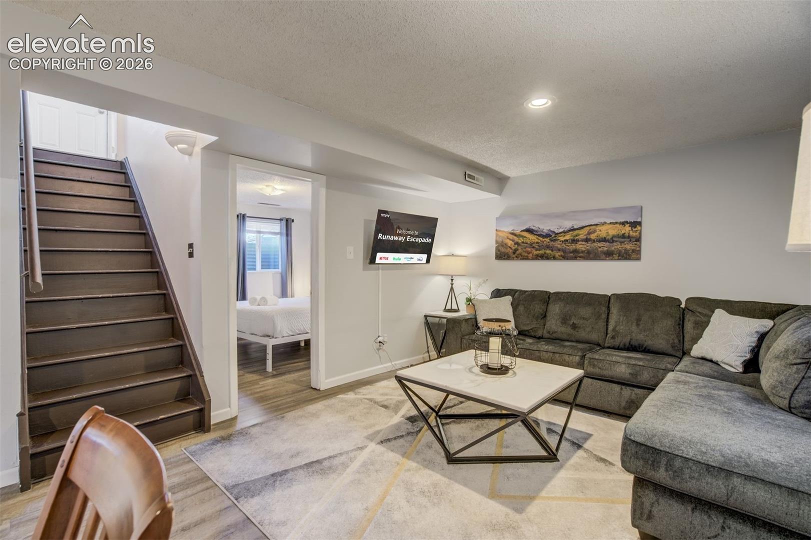 Recessed lighting illuminates the living space featuring light gray walls, wood-finish flooring, and a straight staircase with a wood handrail