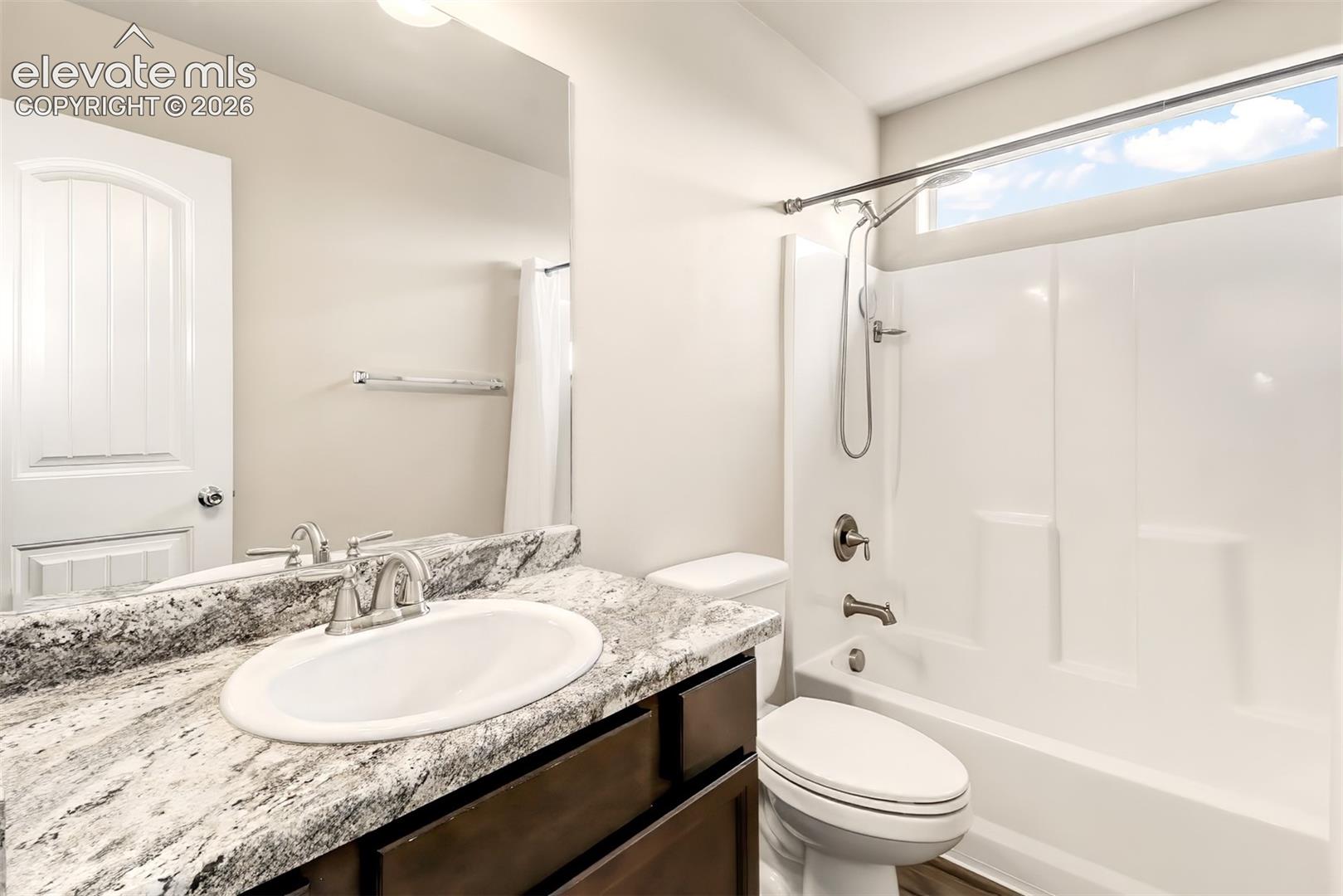 Bathroom featuring a stone-look countertop, undermount sink, dark wood-finish cabinetry, a full-width vanity mirror, and a tub-shower combination with a transom window