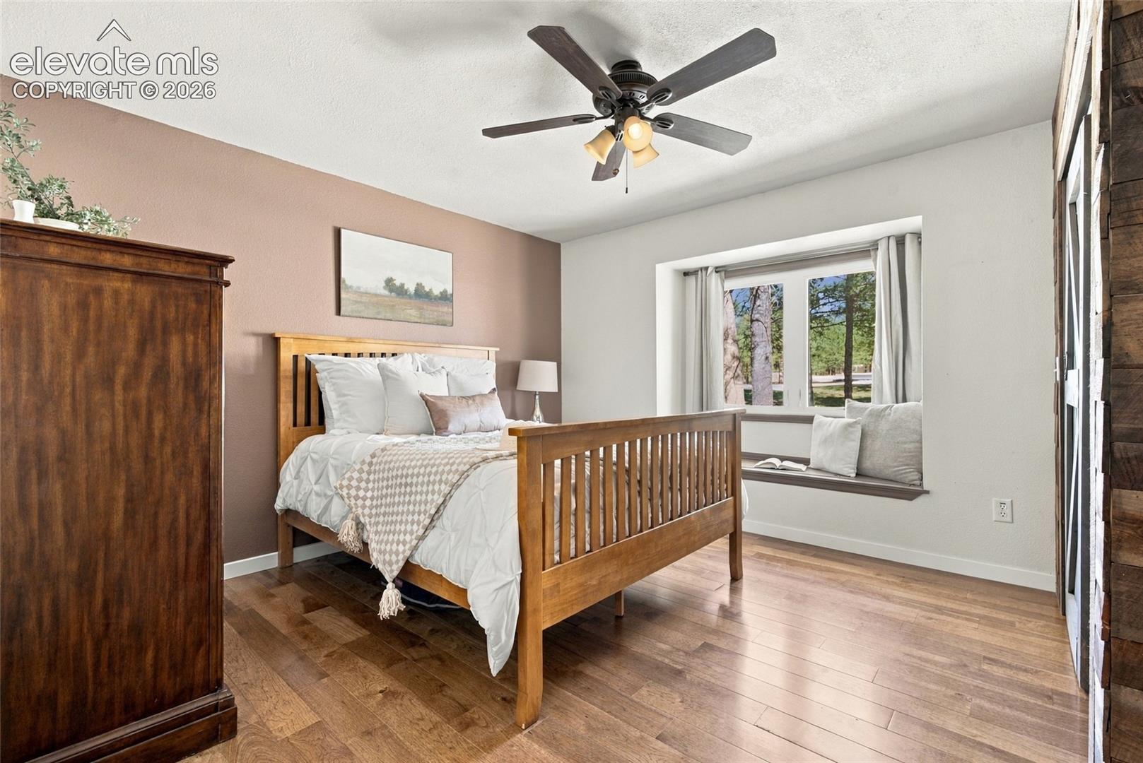 Bedroom with ensuite bathroom, Hardwood flooring, ceiling fan with integrated lighting, window with built-in seating, and a feature wall
