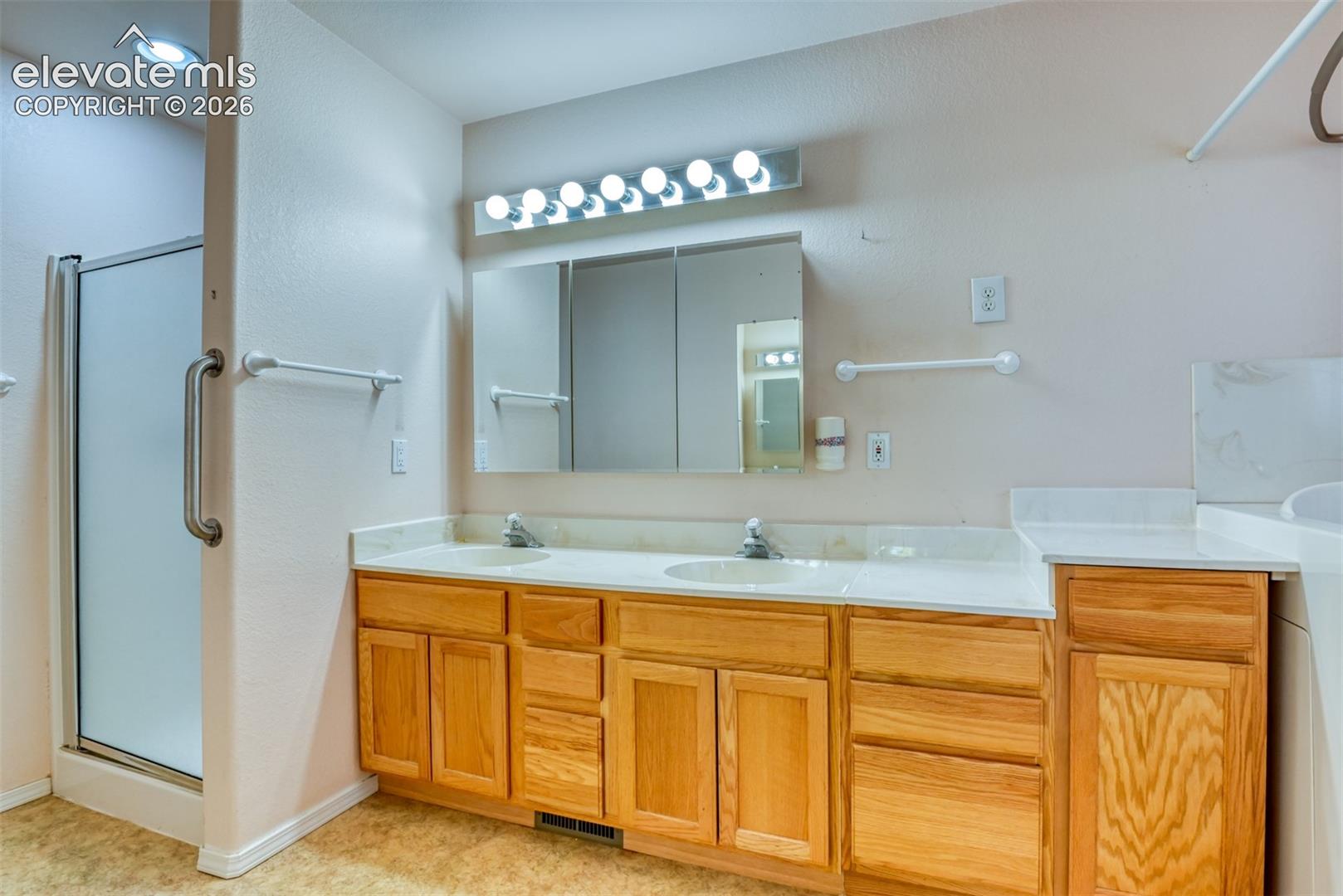 Dual-sink vanity with natural wood-finish cabinetry, solid-surface countertop, integrated mirrored medicine cabinet, and overhead multi-bulb lighting fixture, complemented by an enclosed glass-door shower