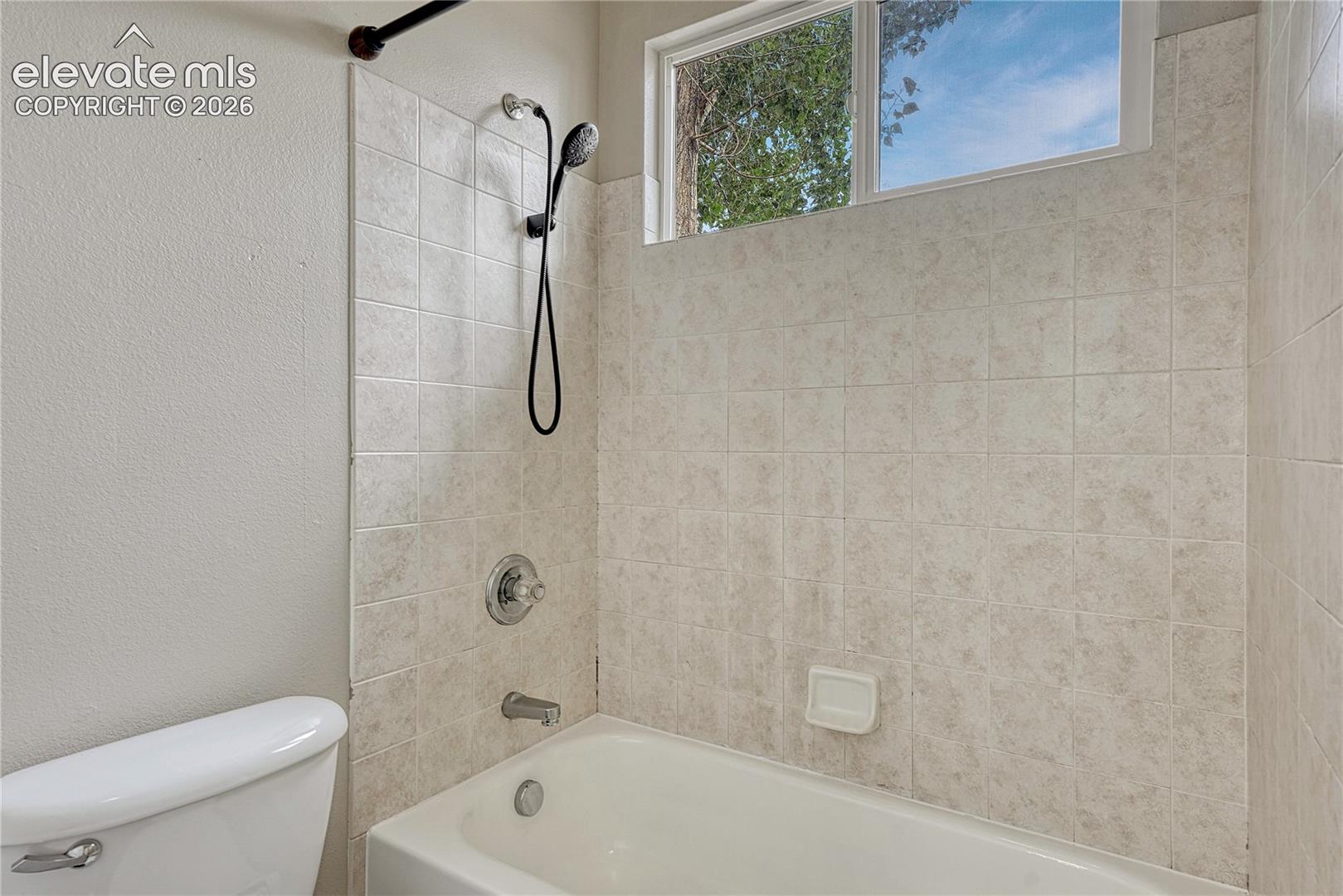 Bathtub and shower combination featuring neutral-toned wall tiling, a handheld shower fixture, and a single-pane slider window