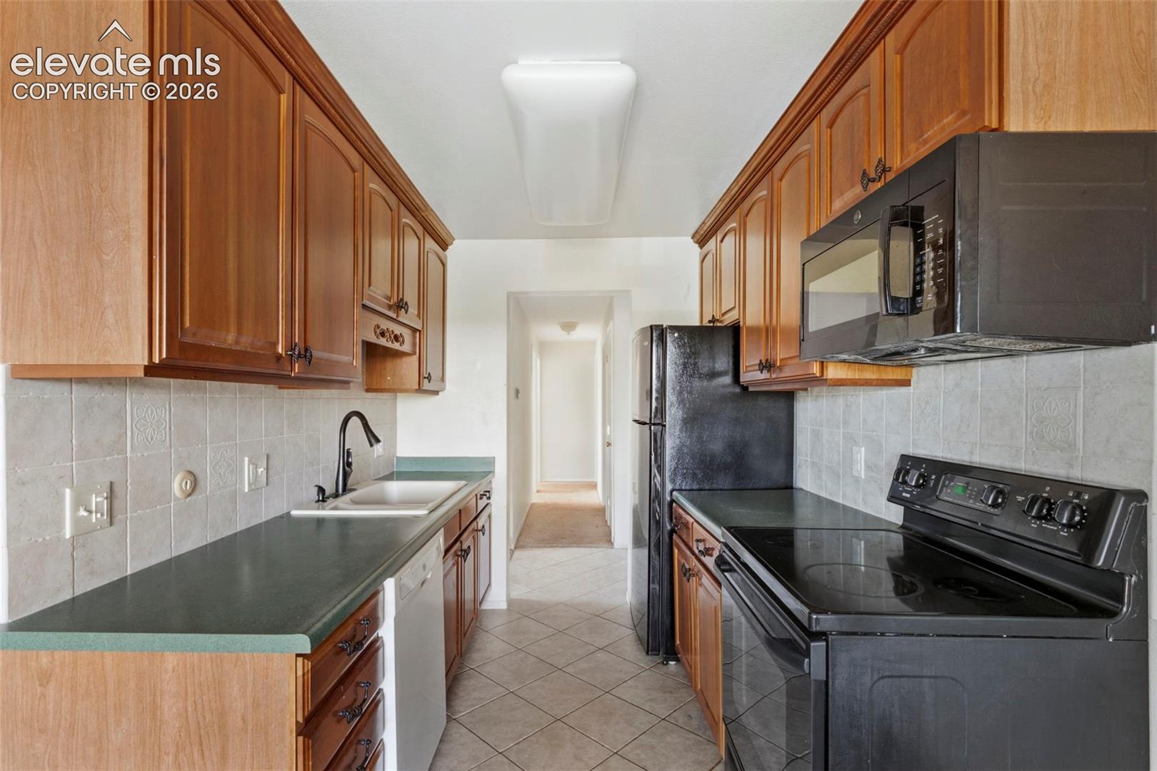 Galley kitchen featuring wood-finish cabinetry, black appliances, a white tile backsplash, and light-toned floor tiling