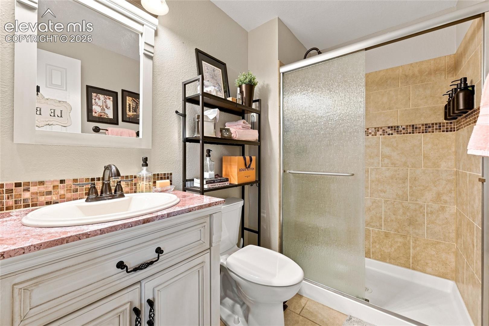 White vanity cabinet with a stone-top counter, undermount sink, framed mirror, and walk-in shower featuring glass sliding doors and tiled walls with a decorative mosaic border