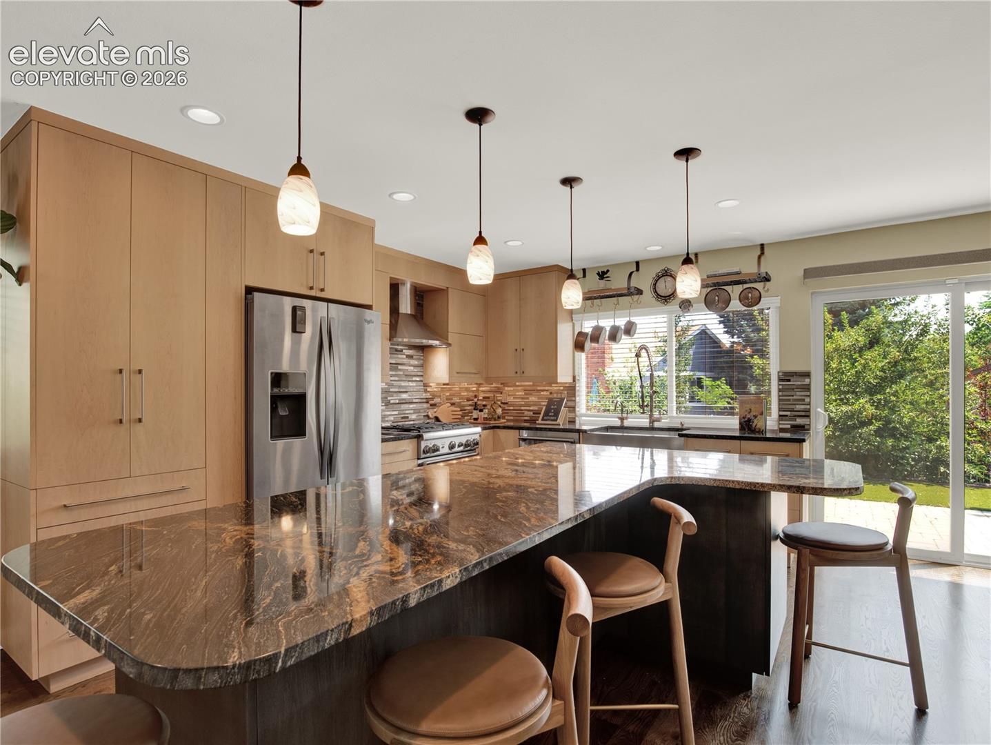 Kitchen featuring flat-panel wood cabinetry, stone countertop island with breakfast bar seating, stainless steel appliances, stacked-stone tile backsplash, and recessed and pendant lighting