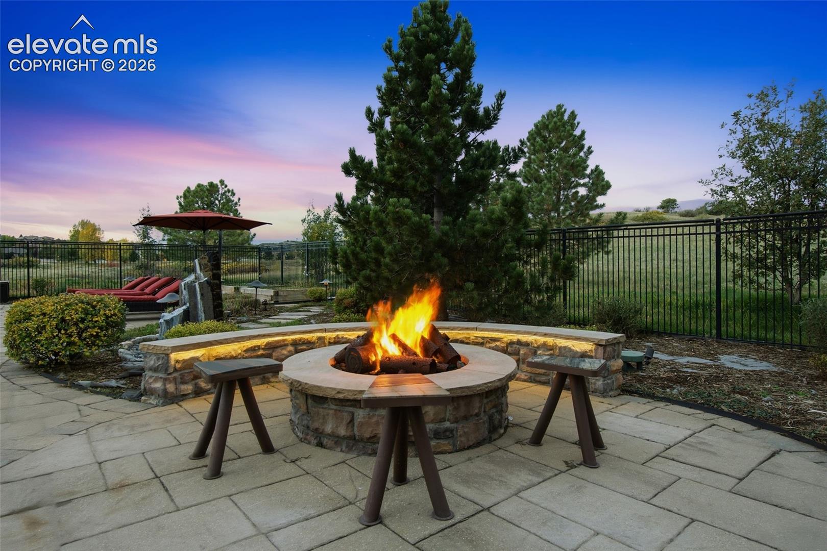 Flagstone patio featuring a circular stone fire pit, curved stone seating wall with integrated lighting, and a metal perimeter fence