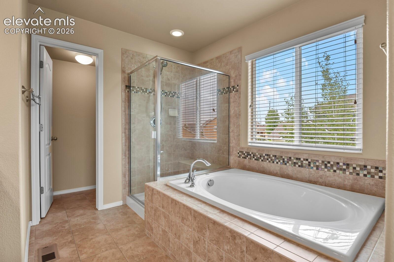 Built-in soaking tub, separate glass-enclosed shower, large window with blinds, tile flooring, and recessed lighting