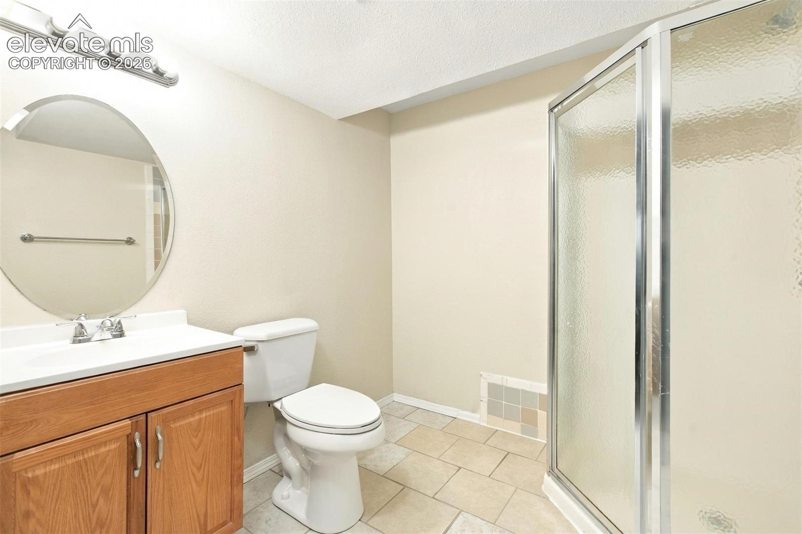 Bathroom featuring a wood-finish vanity with a white countertop, oval wall-mounted mirror, ceramic tile flooring, and a glass-enclosed shower stall