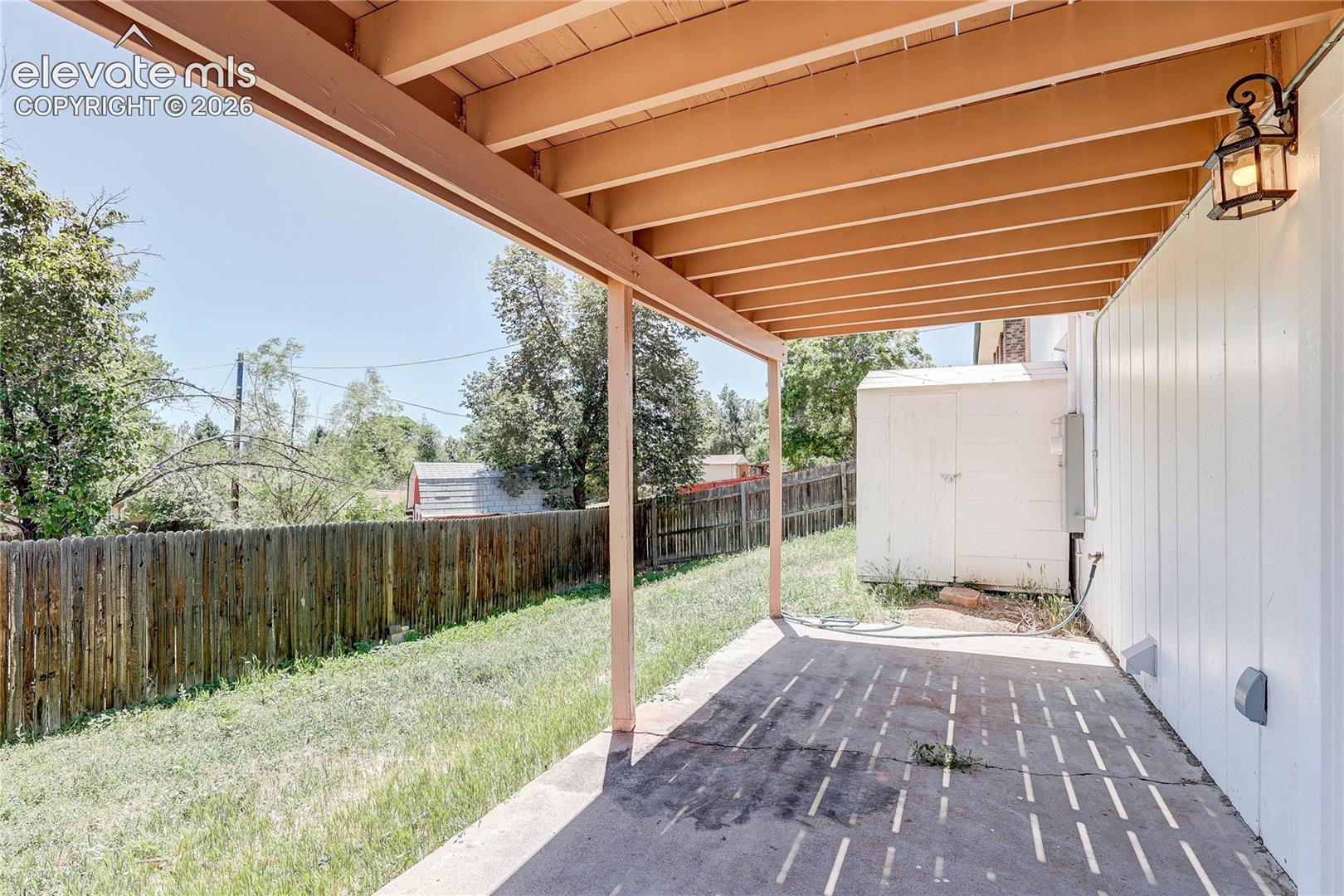 Covered concrete patio featuring an exposed timber beam ceiling, wall-mounted lantern-style lighting, and vertical siding, complemented by a fenced backyard with an exterior storage structure