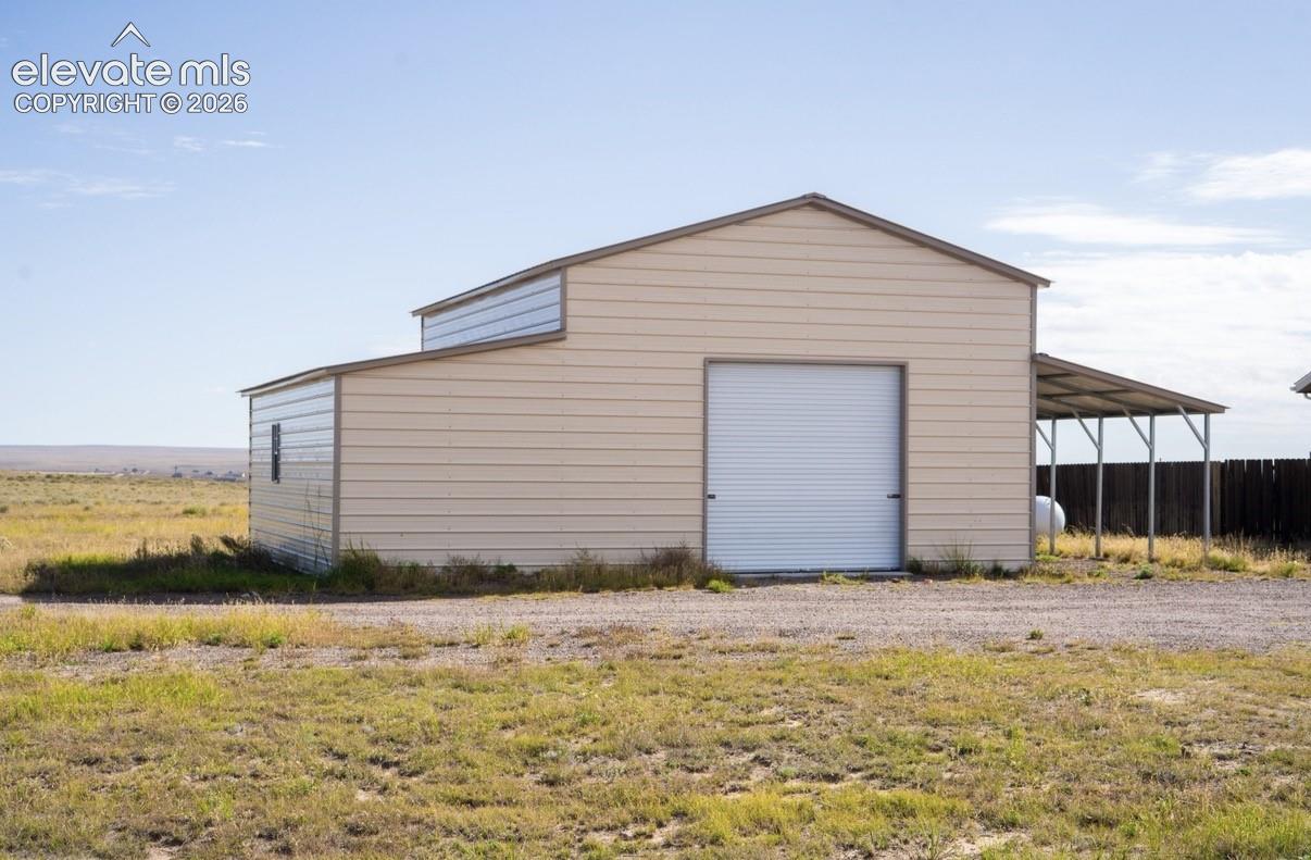 Detached metal outbuilding featuring high-profile roll-up door, horizontal siding, raised center roofline, and an attached side carport