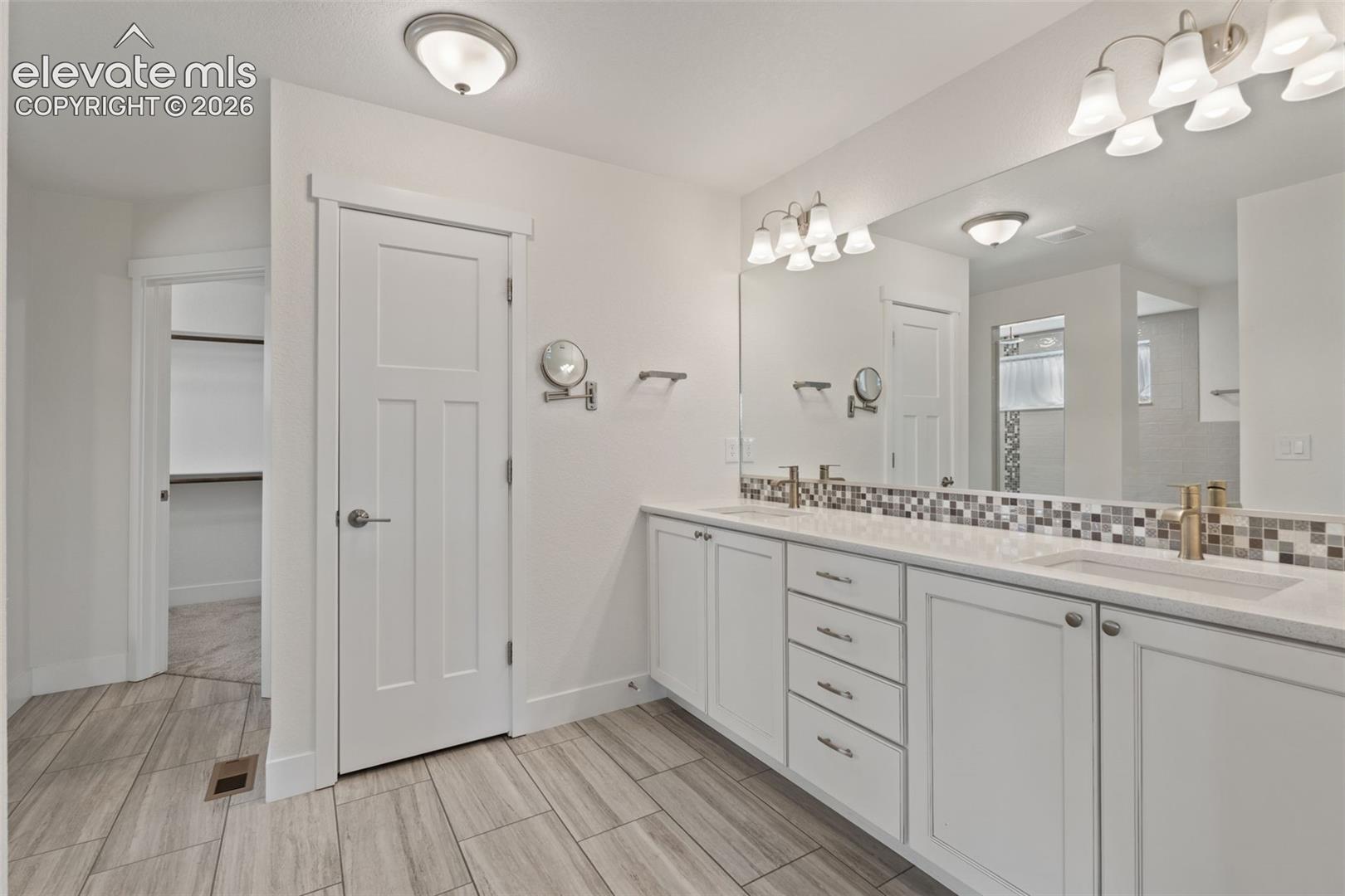 Dual vanity with undermount sinks, wood-finish tile flooring, recessed built-in shelving, white shaker-style cabinetry, and a full-width mirror