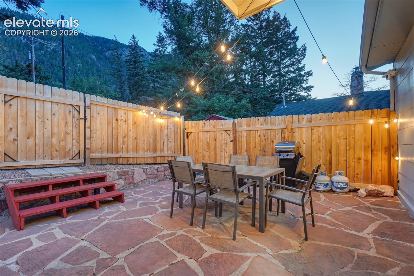 Expansive flagstone patio with a privacy fence, stone retaining wall, and string lighting