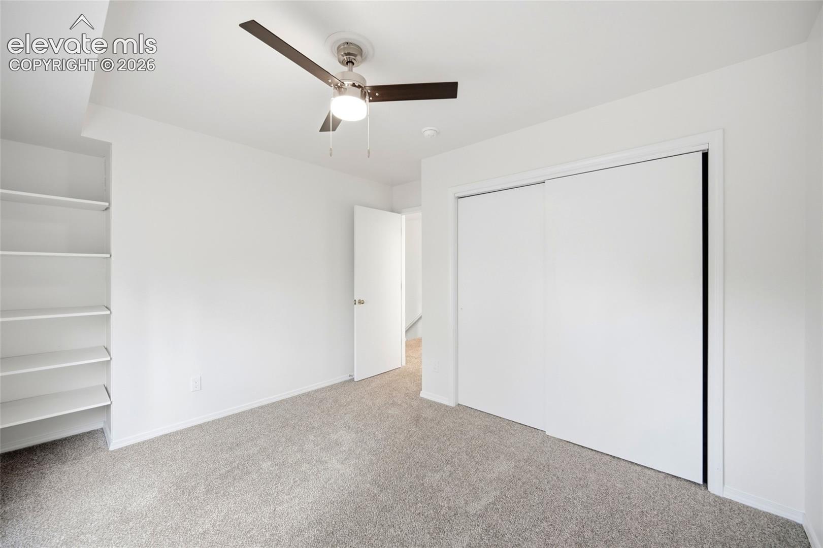 Interior space featuring neutral carpeting, a dual-panel sliding closet, built-in shelving, and a ceiling-mounted light fixture with a three-blade fan