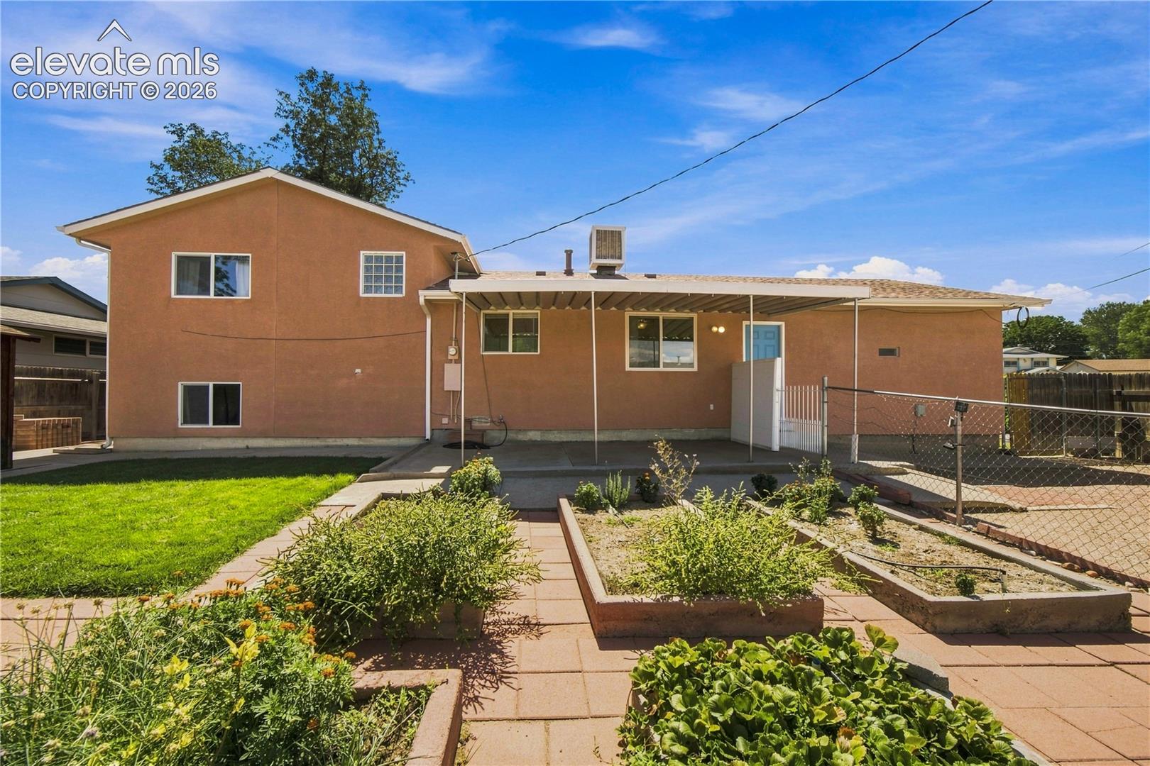 Rear exterior with stucco finish, a covered patio, a fenced area, a grass lawn, and raised garden beds