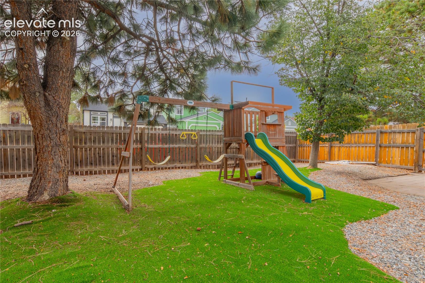 Enclosed backyard featuring artificial turf, mature trees, a wooden privacy fence, and a gravel pathway